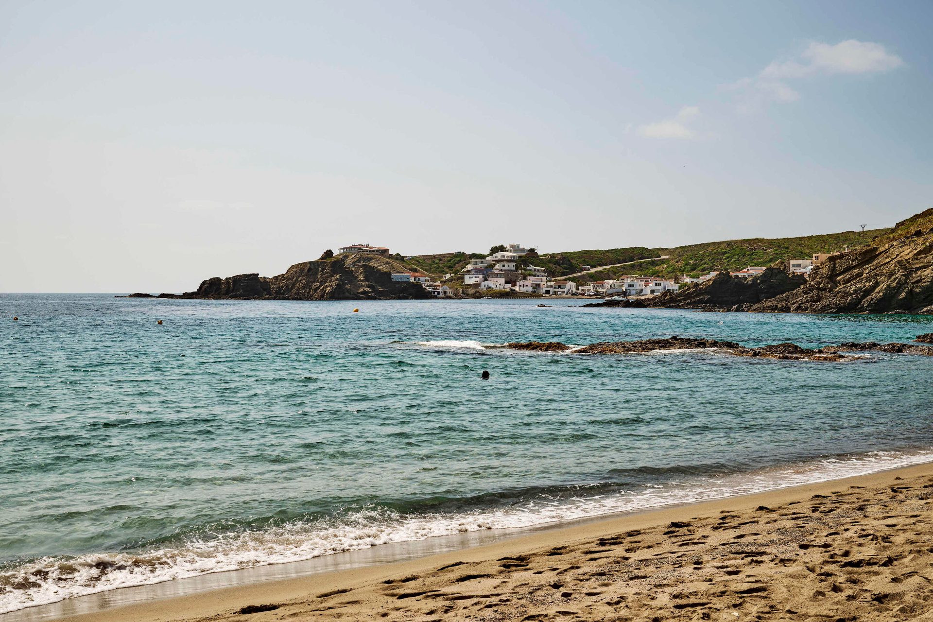 Sandy beach, turquoise sea, and a village on rocky, green hills by the coast.