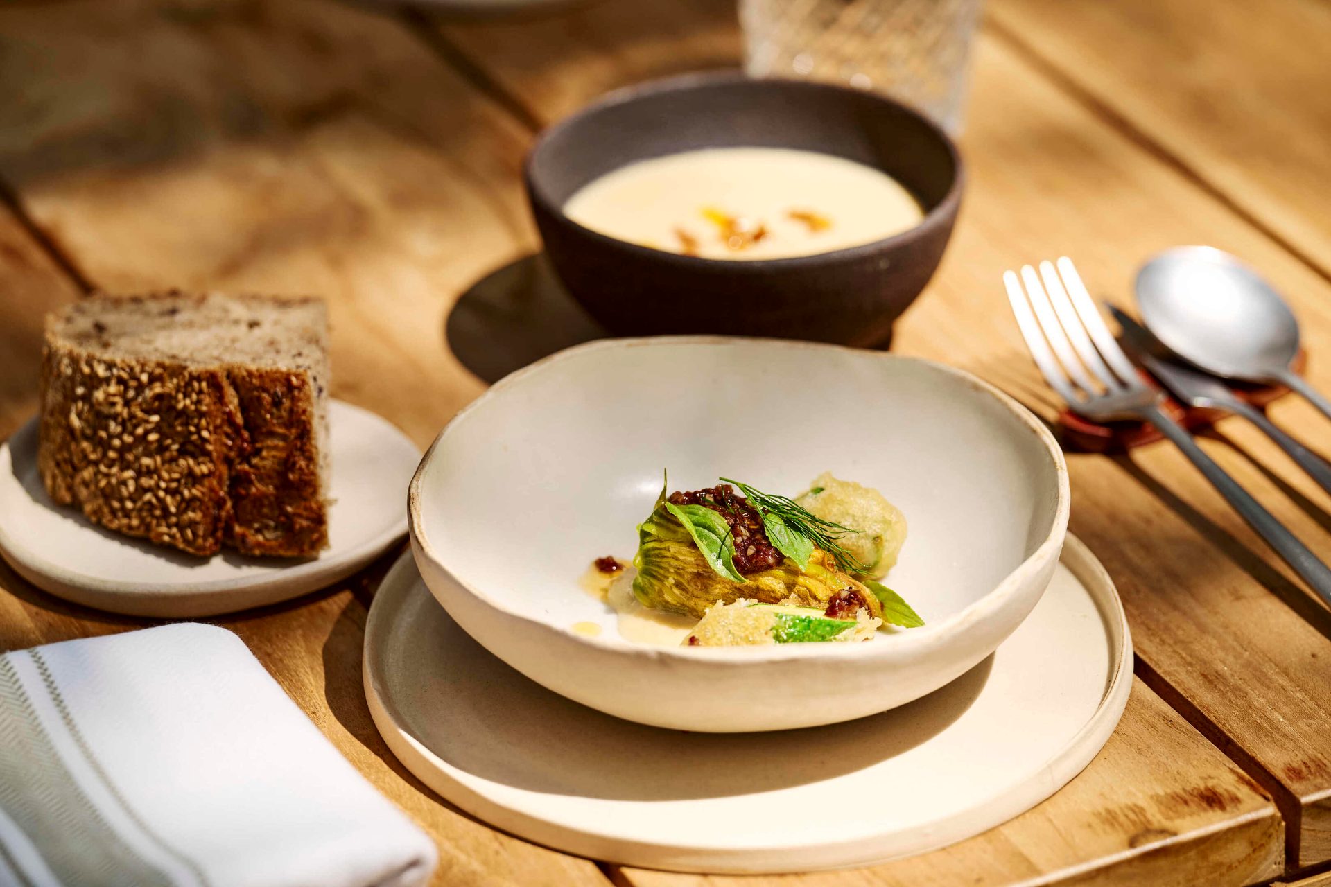 Gourmet meal with soup, stuffed squash blossom, and bread on a wooden table outdoors.