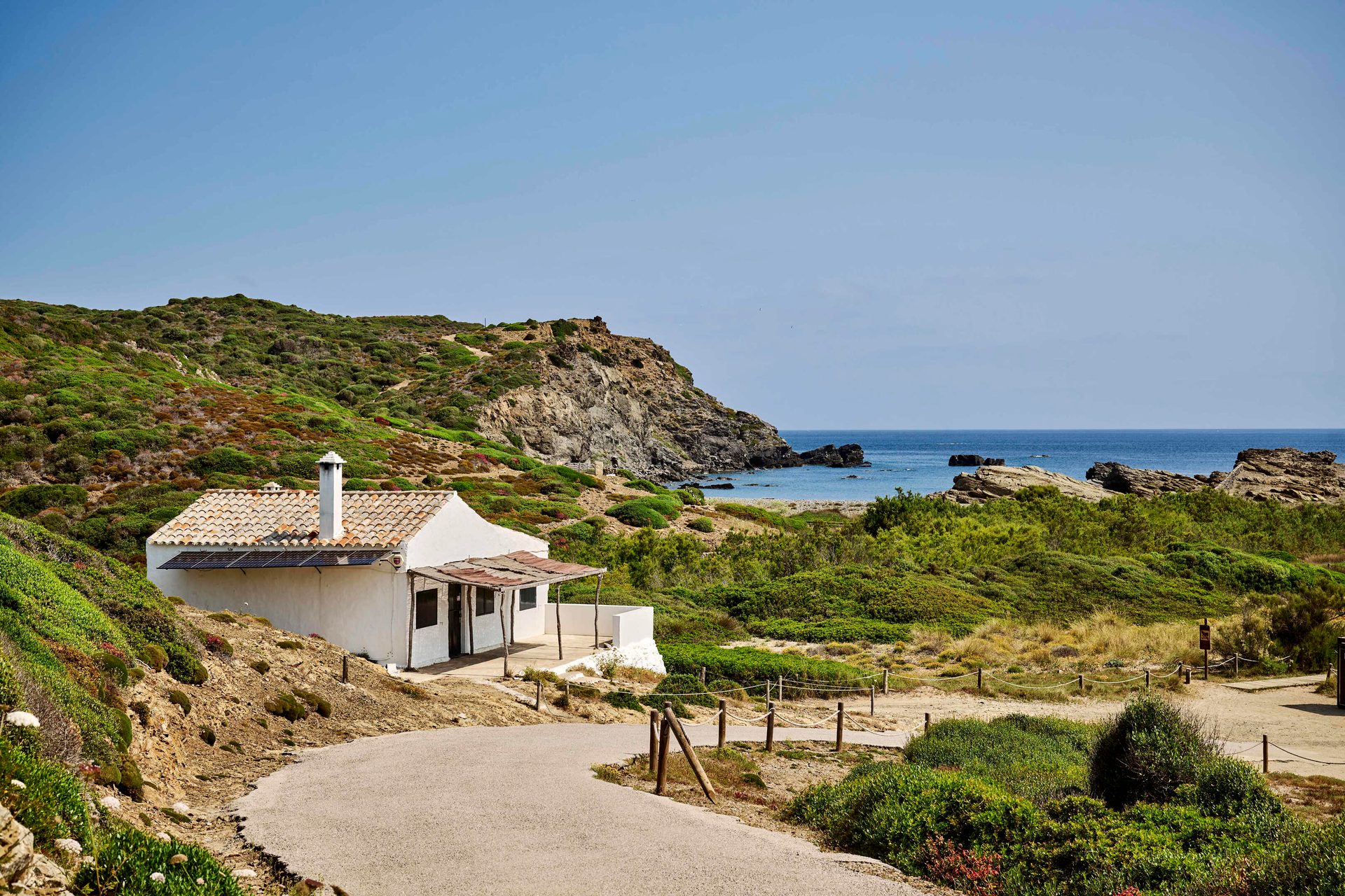 A white house with a tiled roof nestled among green hills, overlooking a rocky coastline and the blue sea.