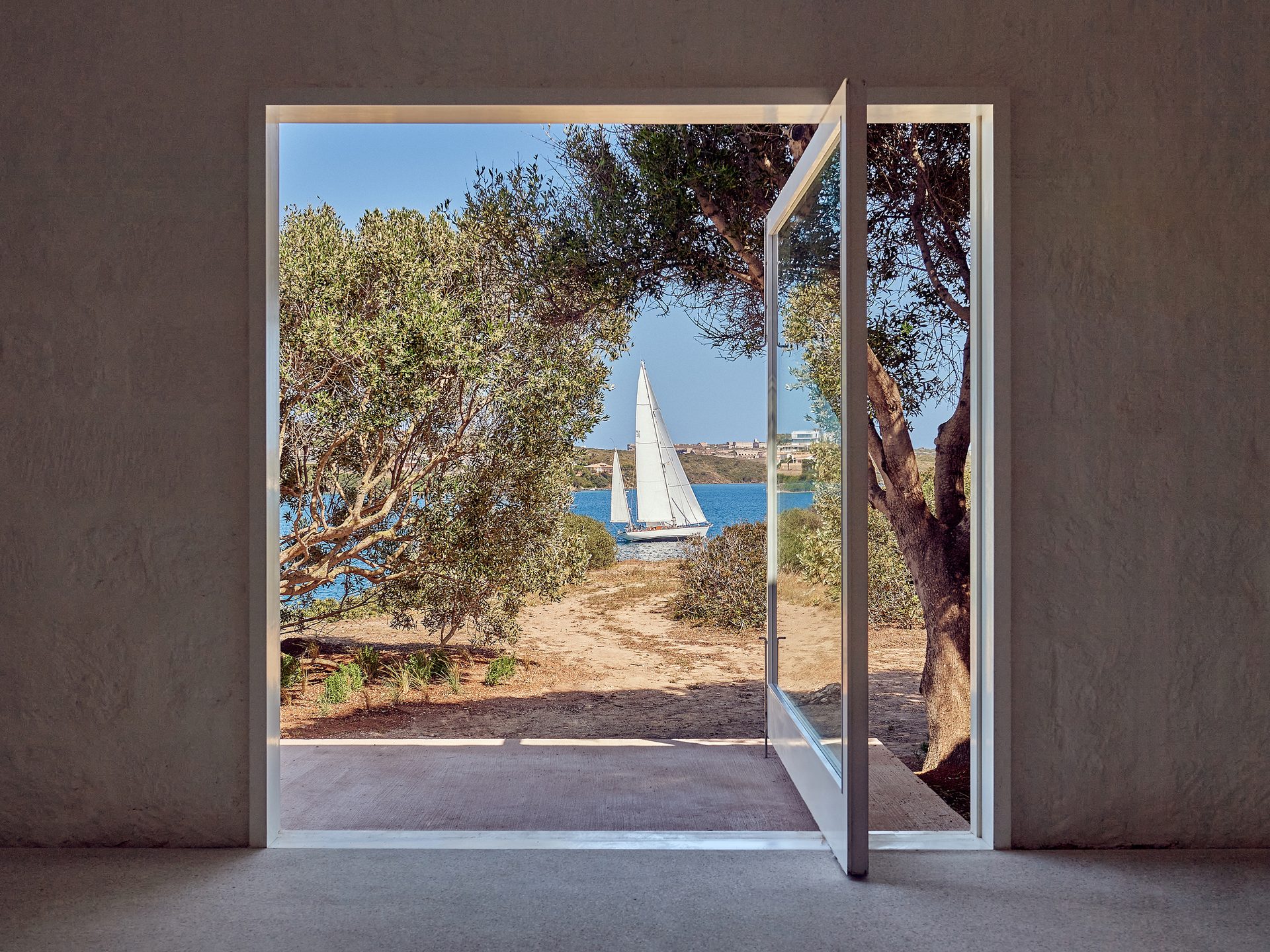 Sailboat on blue water seen through open door and trees.