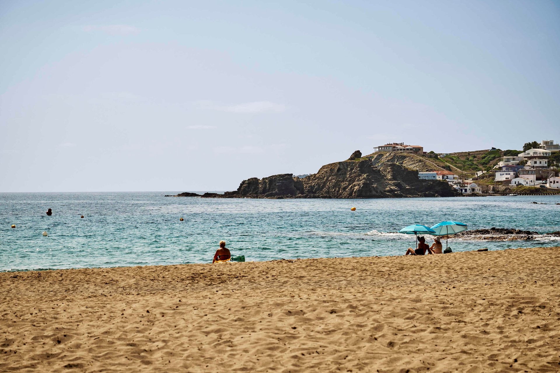 Sunny beach with people, blue sea, and a village on a rocky coastline.