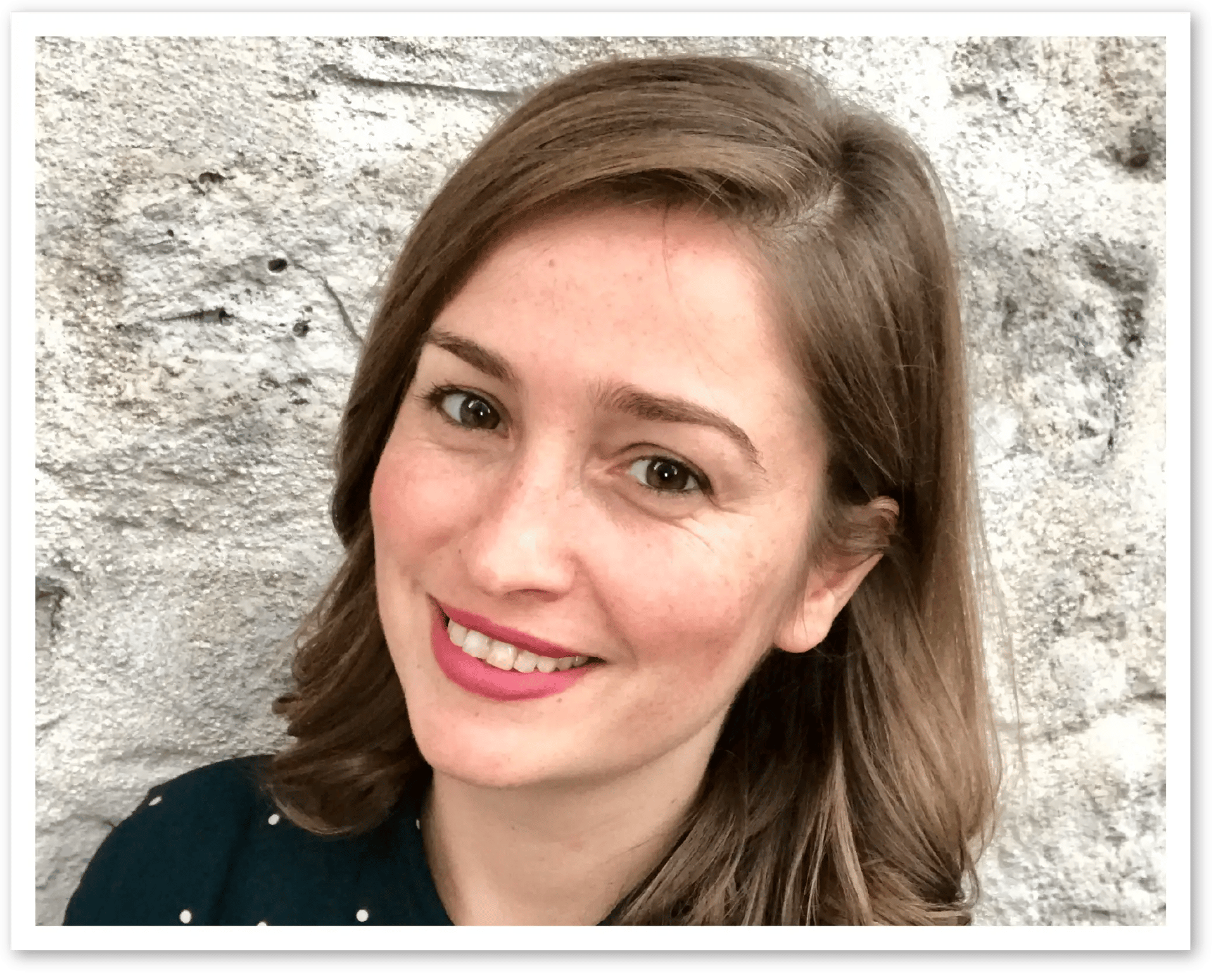 Headshot of a smiling woman with brown hair and a dark polka dot top against a stone wall.