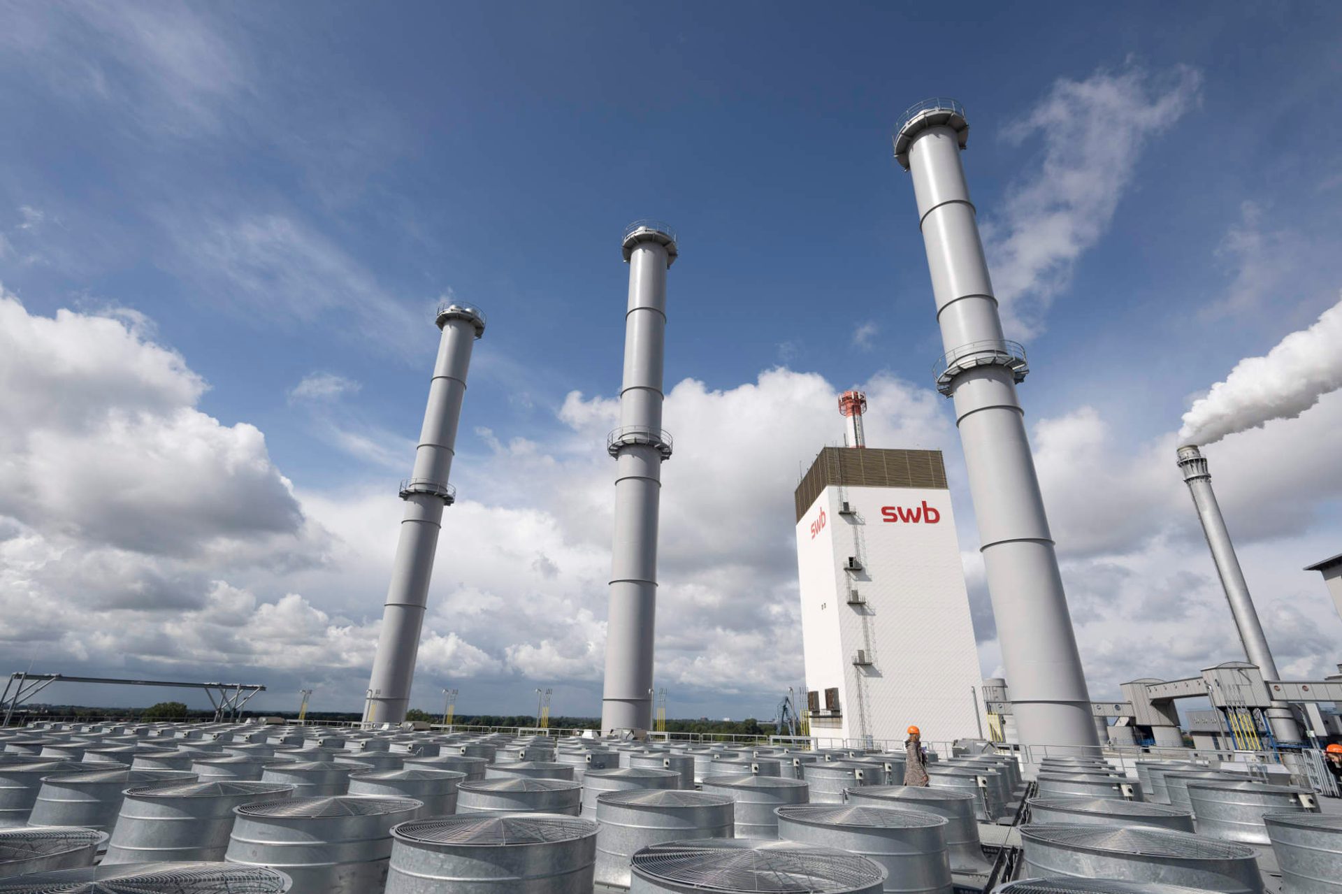 Power plant with tall chimneys, an swb building, and rooftop cooling units under a cloudy blue sky.