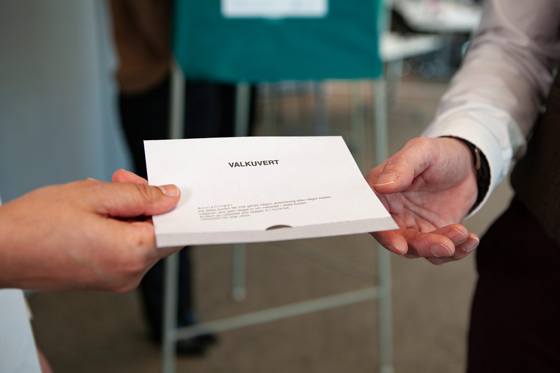 Two hands exchanging a white ballot envelope labeled "VALKUVERT" at a polling station.