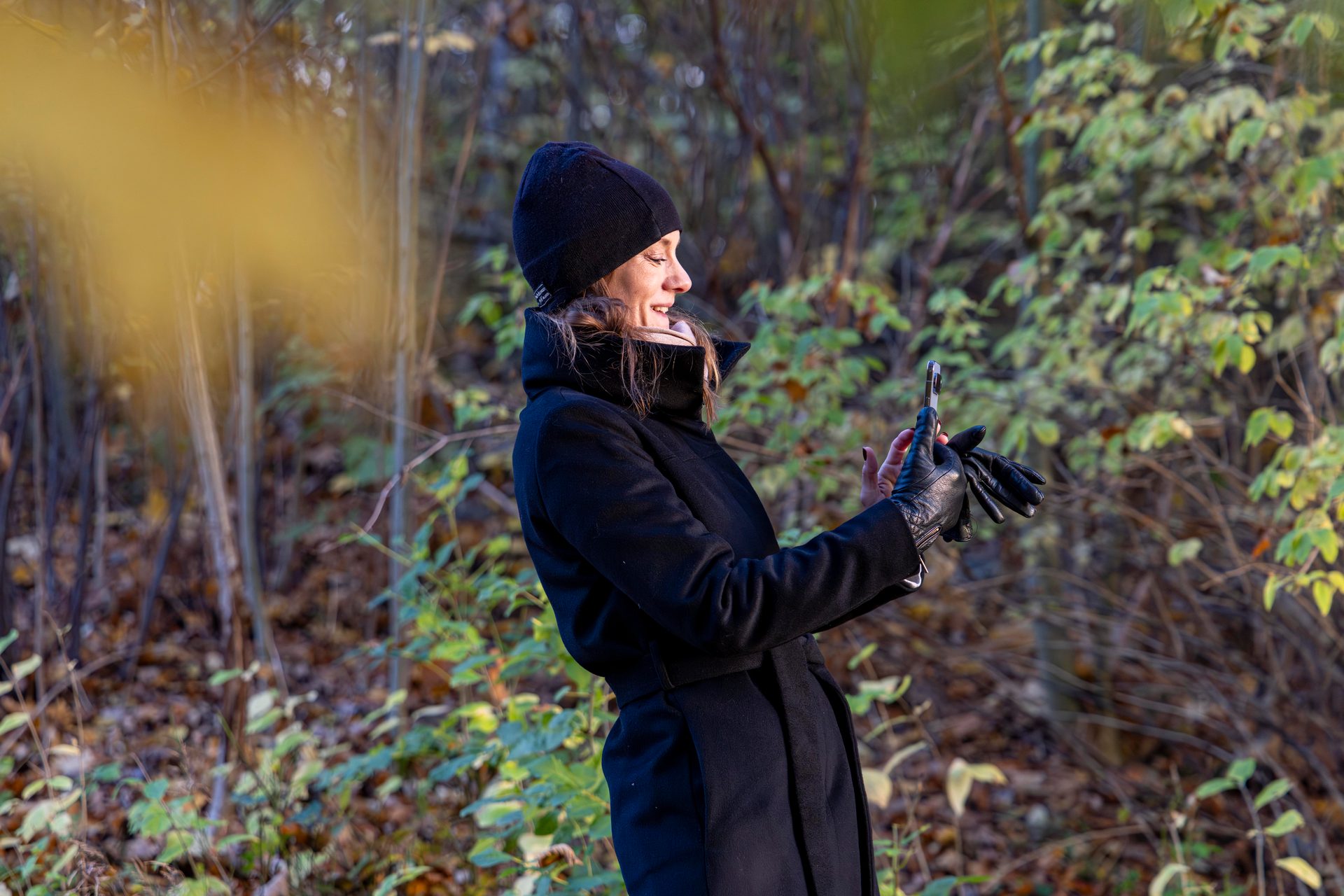 Smiling woman in a black coat and beanie holds a phone and gloves in a forest.