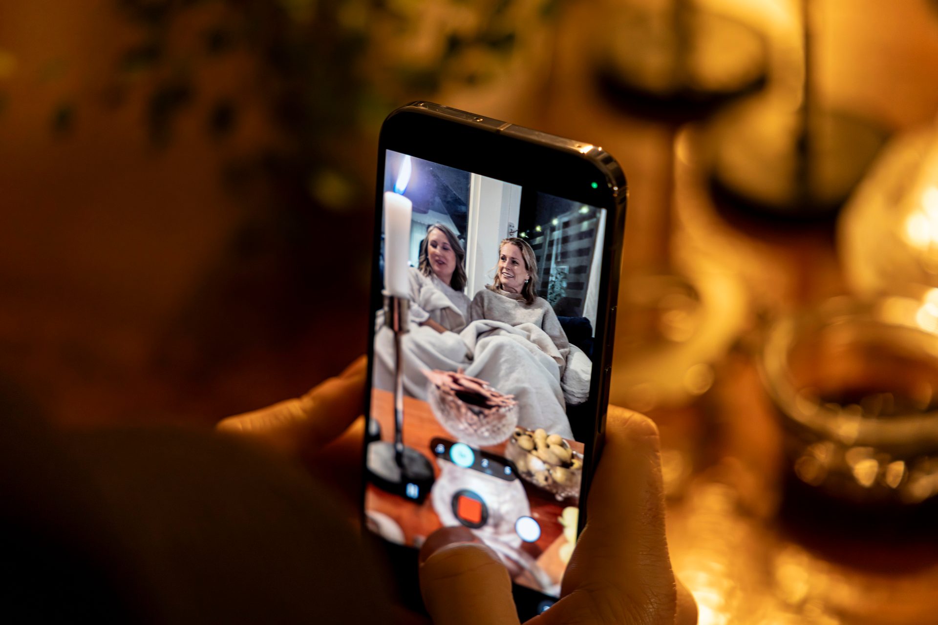 Hands hold a smartphone displaying two women on its screen, with a lit candle nearby.