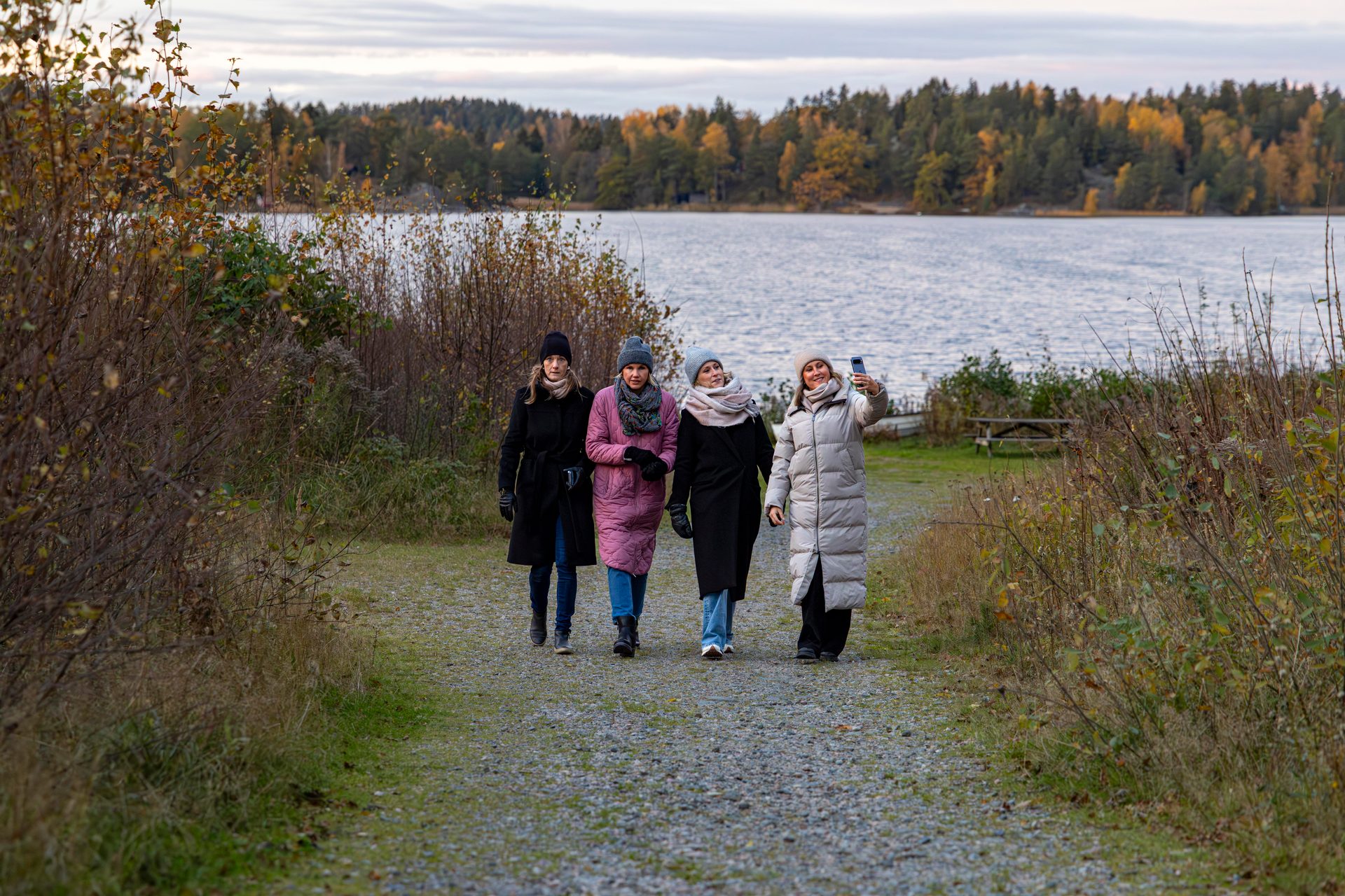 Four women walking on a path by a lake, one taking a selfie with a phone.
