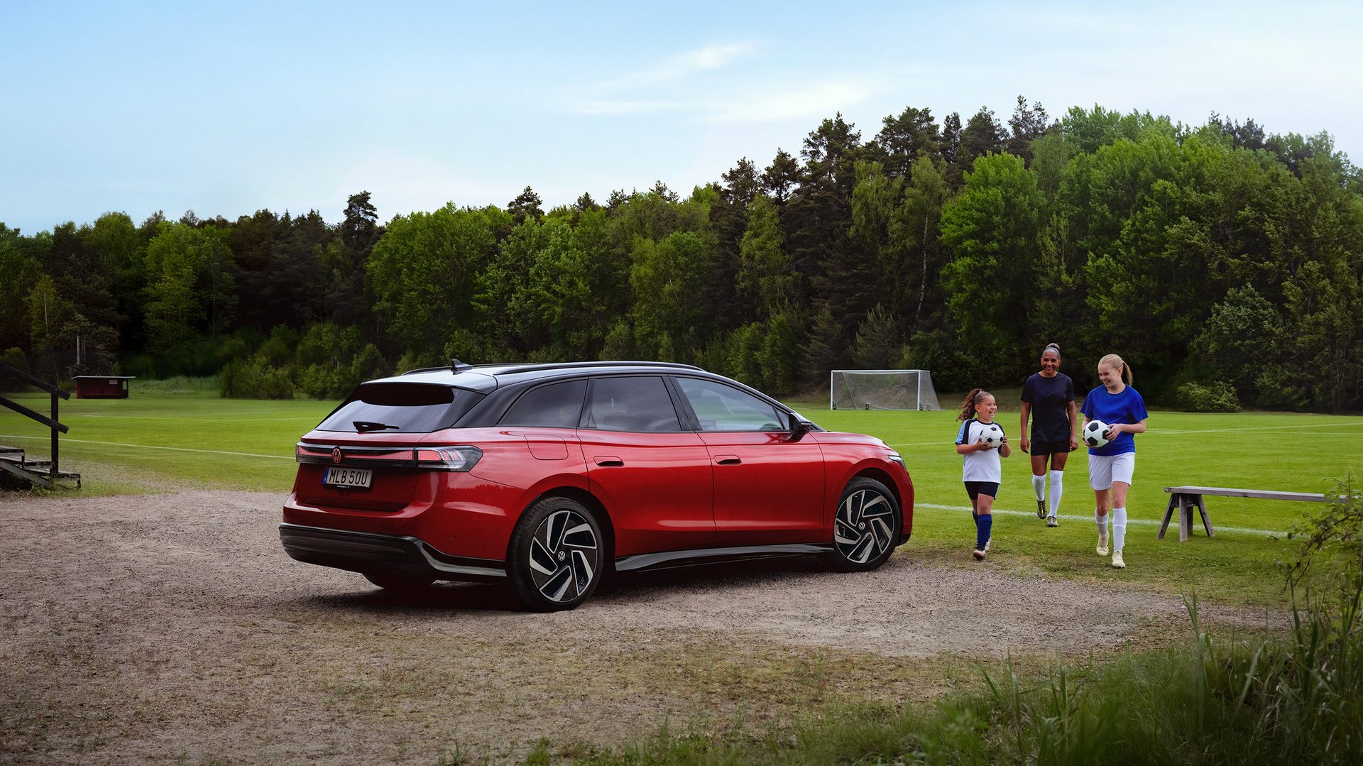 Red car parked at a soccer field with three soccer players holding balls.