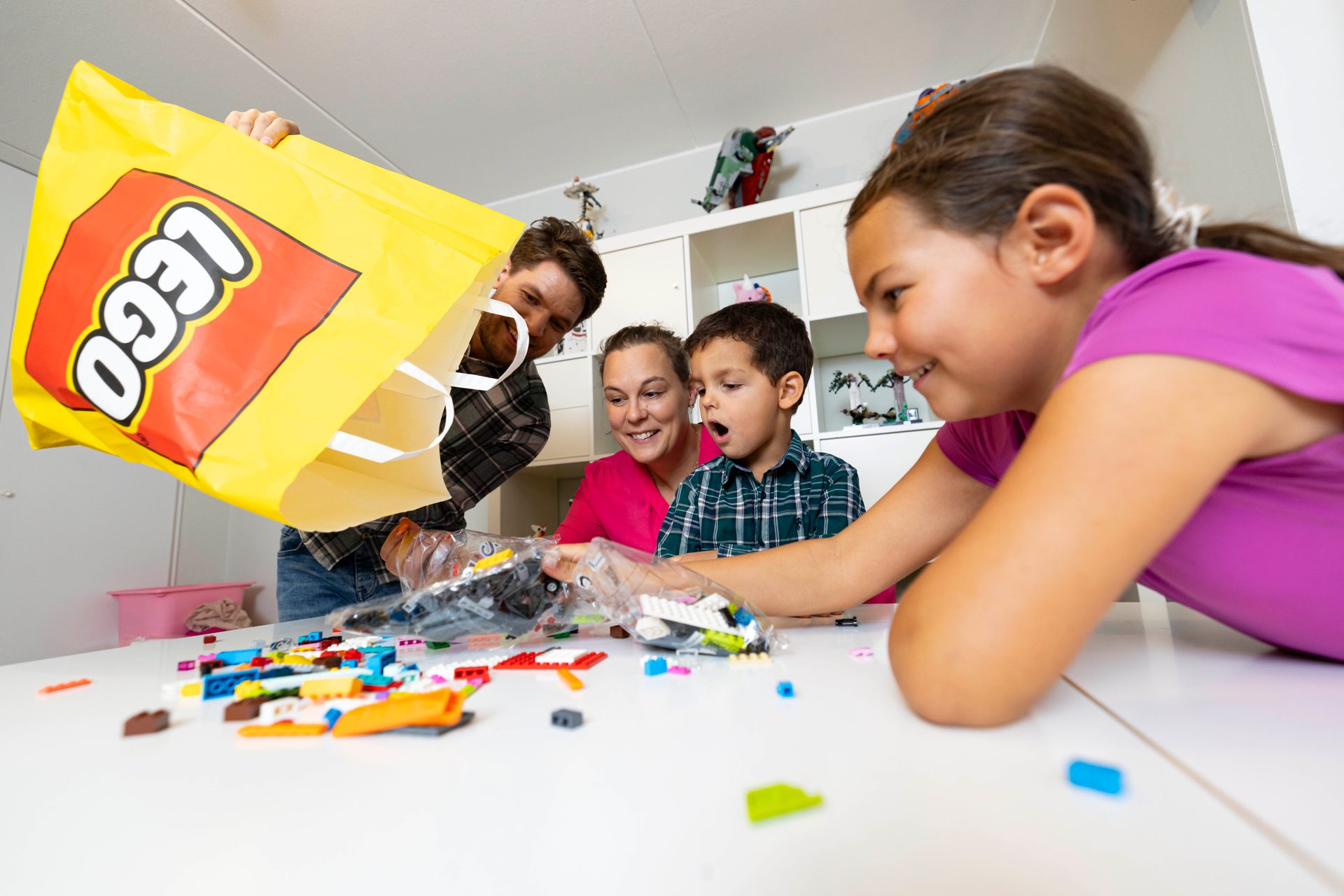 A family excitedly empties a large yellow LEGO bag, spilling colorful bricks onto a table.