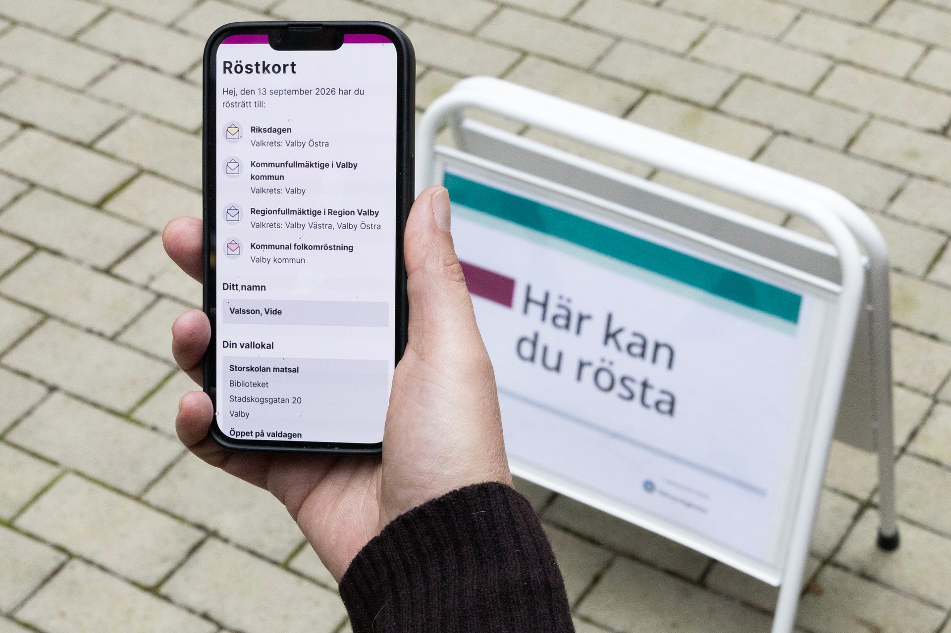 A hand holds a smartphone displaying a digital voting card, with a "Here you can vote" sign in the background.