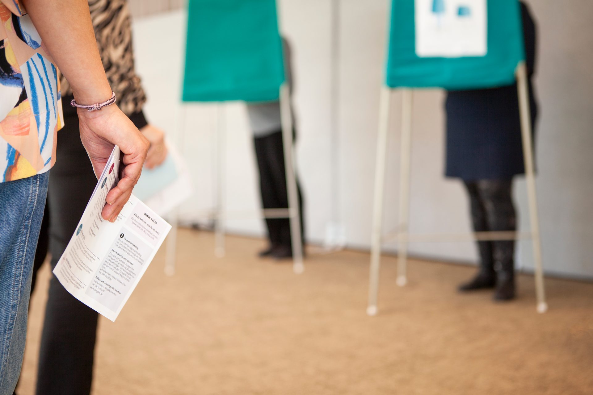 Close-up of a person's hand holding voting instructions at a polling station with blurred booths.