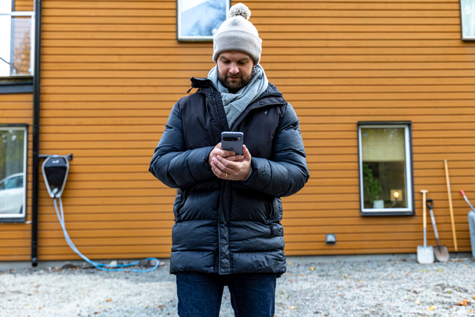 A man in winter gear looks at his phone in front of an orange house.