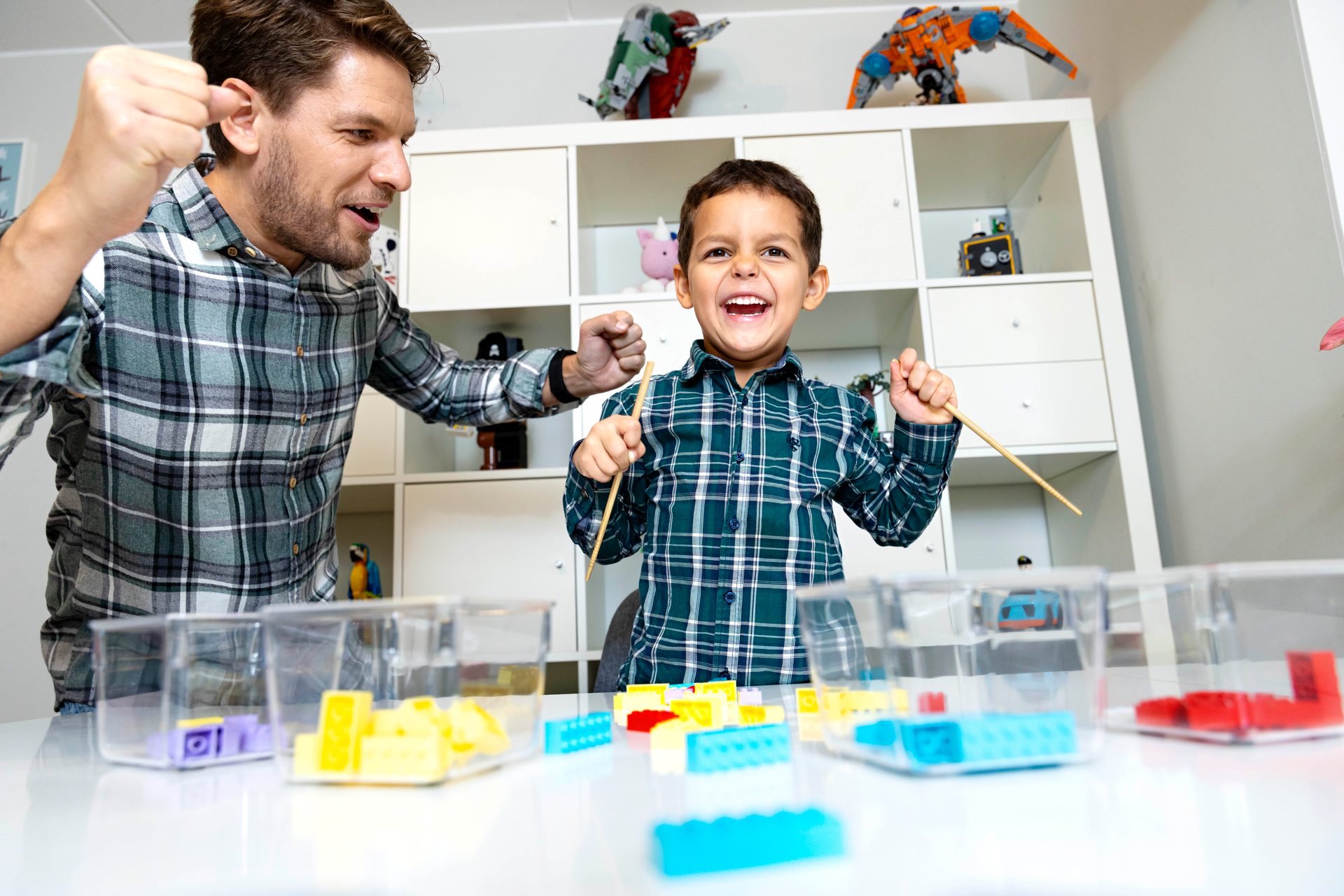 Joyful father and son in plaid shirts enthusiastically playing with colorful building blocks; son holds a stick.