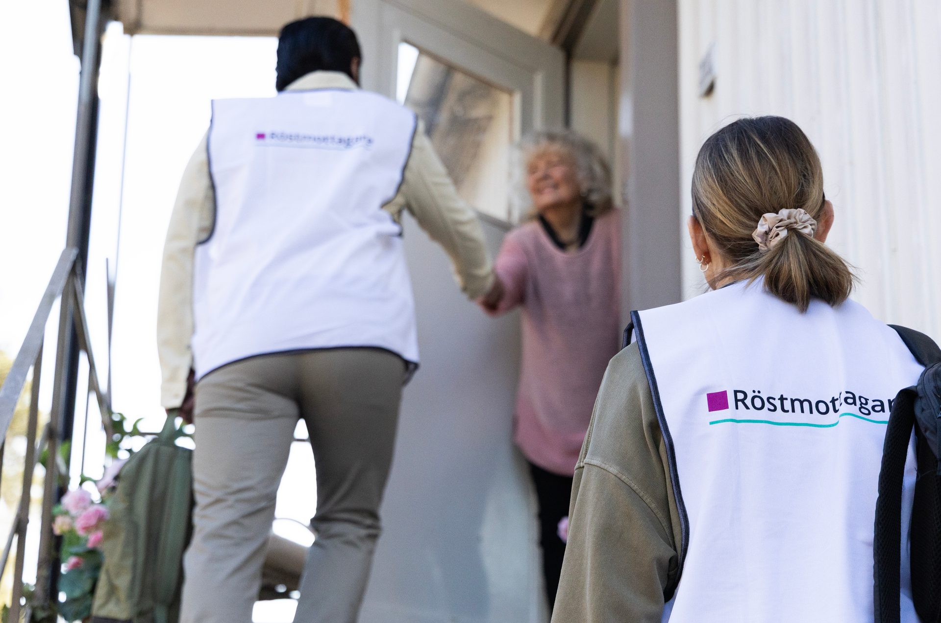 Two people in "Röstmottagare" vests assist an elderly woman at her doorstep, holding hands.