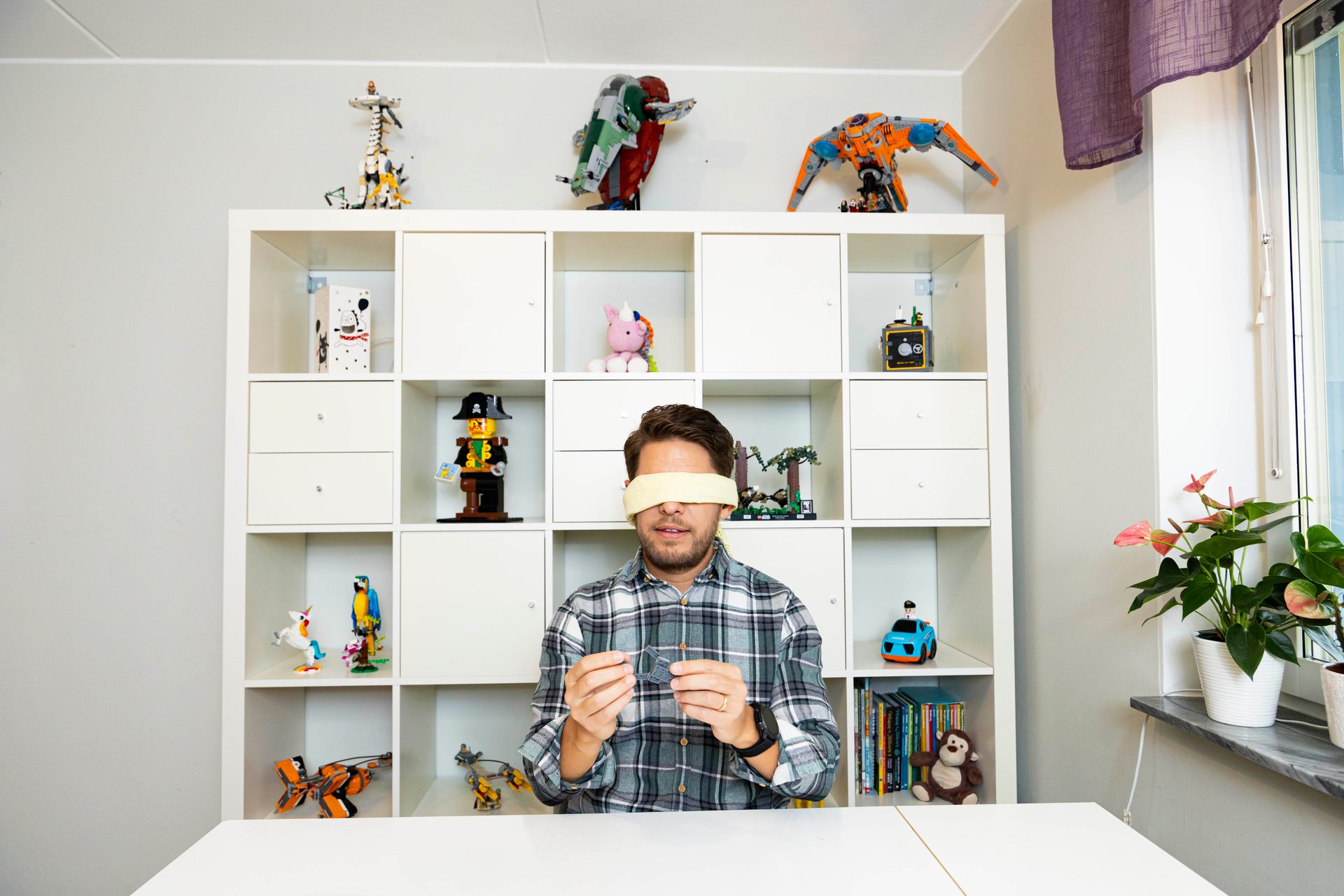 Blindfolded man holding Lego pieces in front of a shelf filled with Lego models.