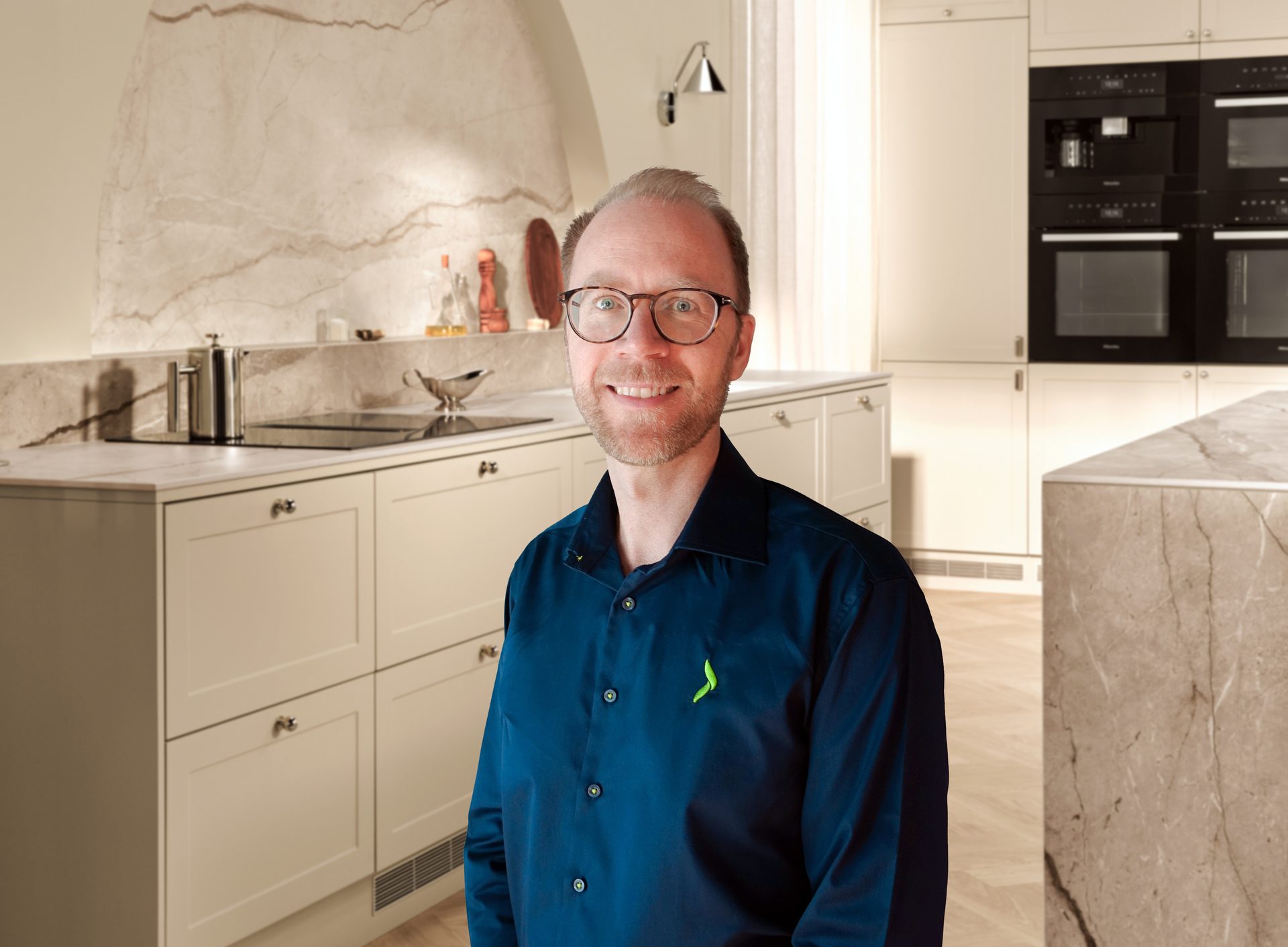 A smiling man in glasses and a dark blue shirt stands in a modern kitchen with marble accents.