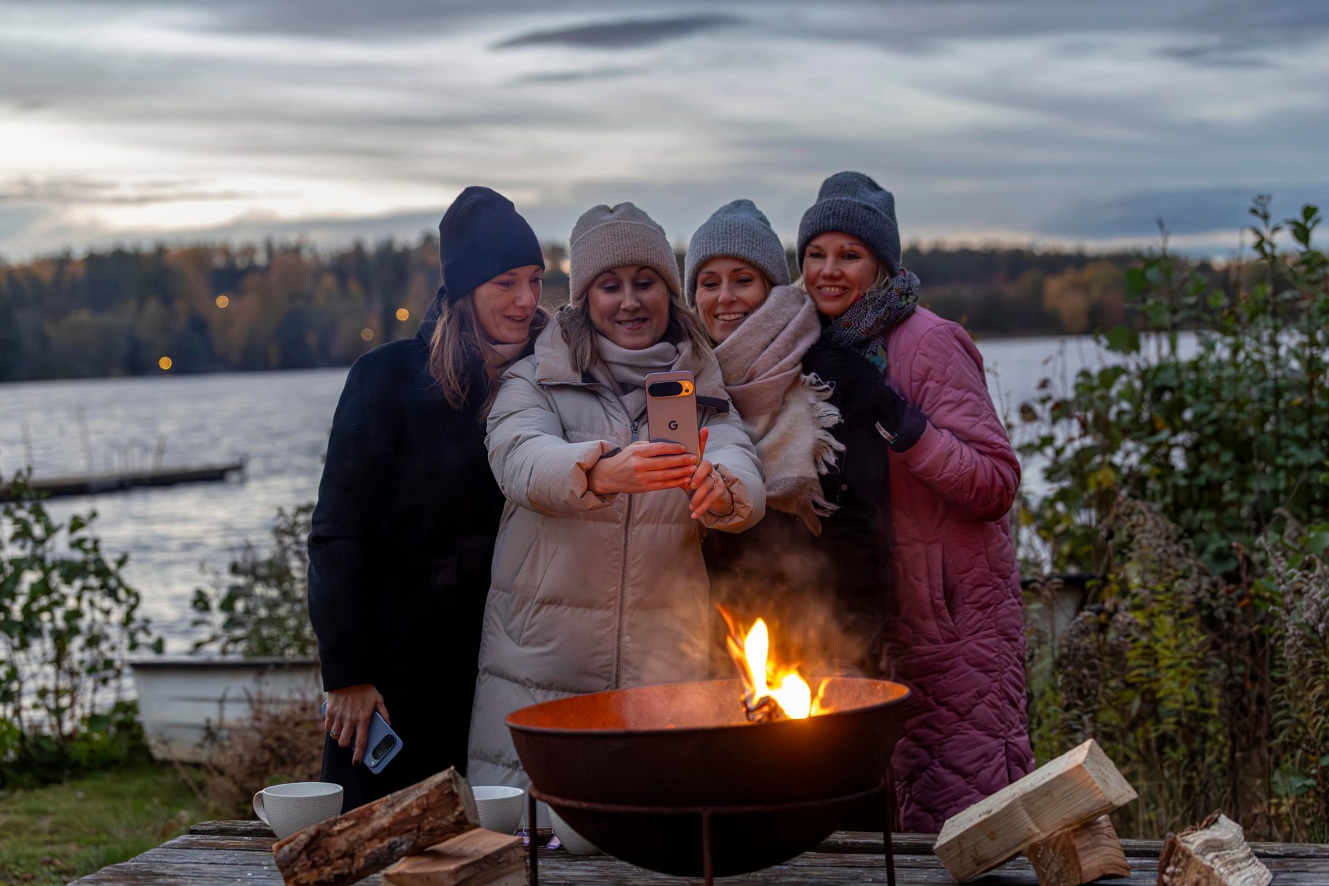 Four women in winter clothes taking a selfie by a lake with a fire pit at dusk.