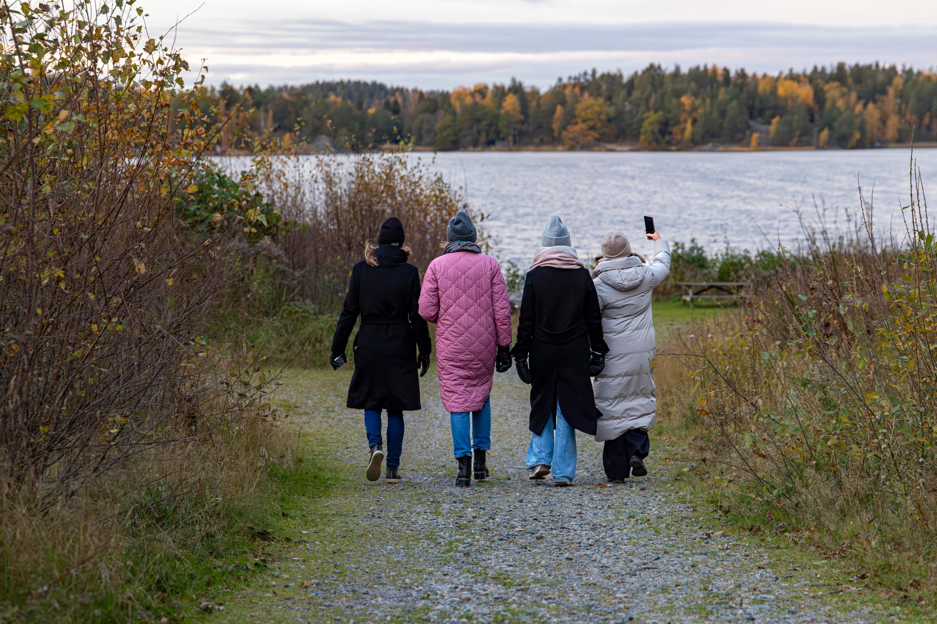 Four people walk on a path towards a lake with autumn trees, one taking a photo.