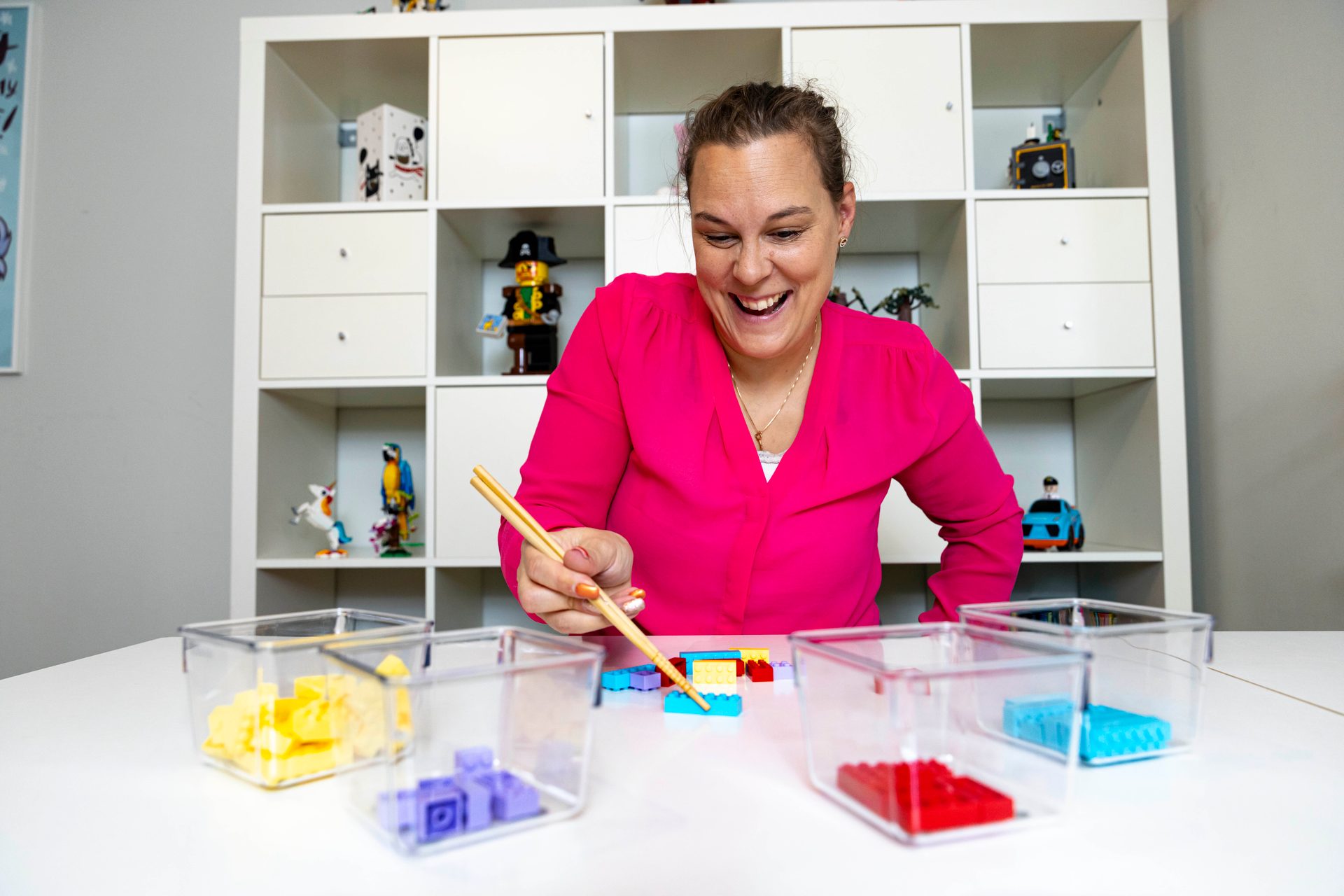 A smiling woman in a pink shirt uses chopsticks to pick up colorful Lego bricks.