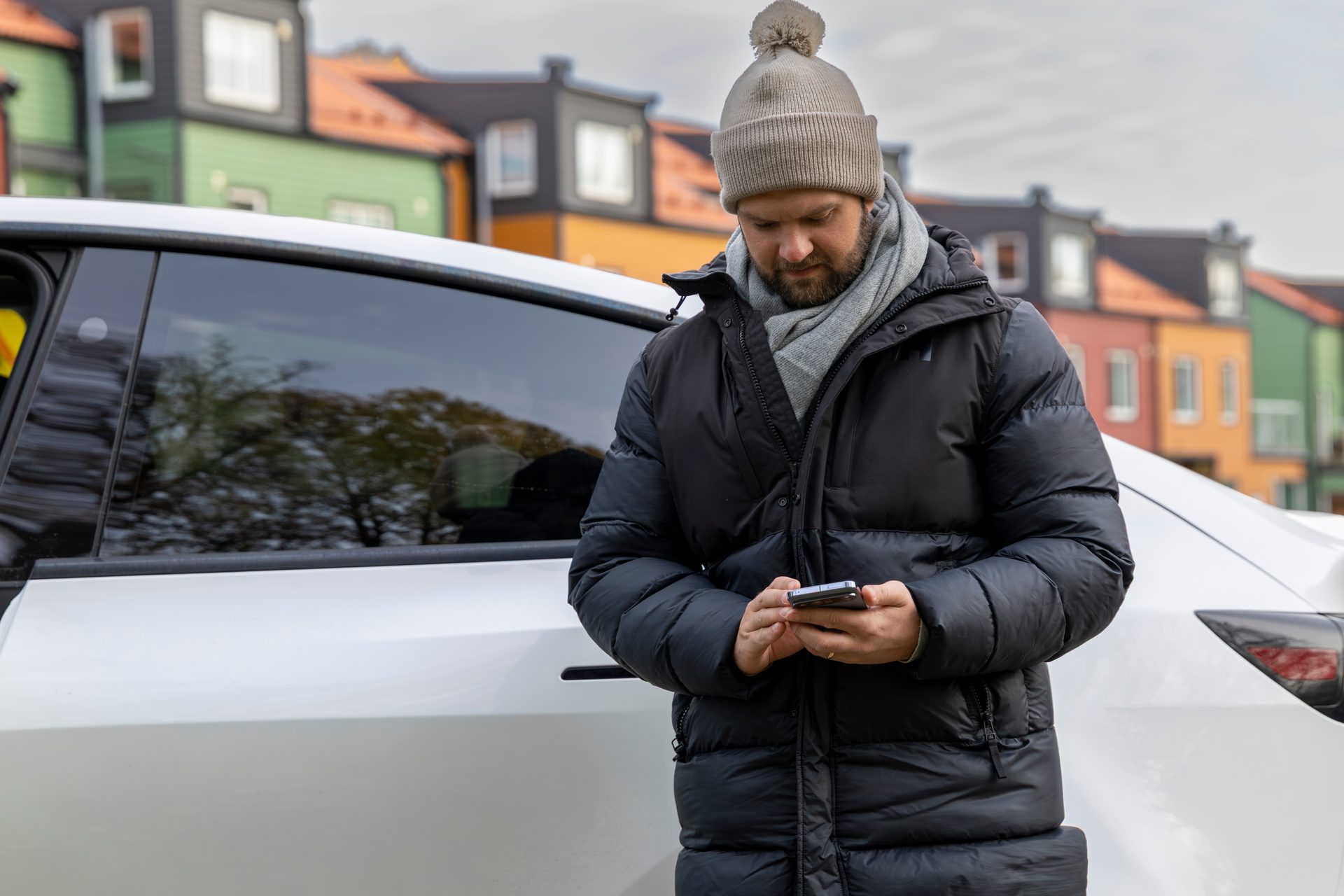 Man in winter clothes looks at phone by white car, colorful houses blurred in background.