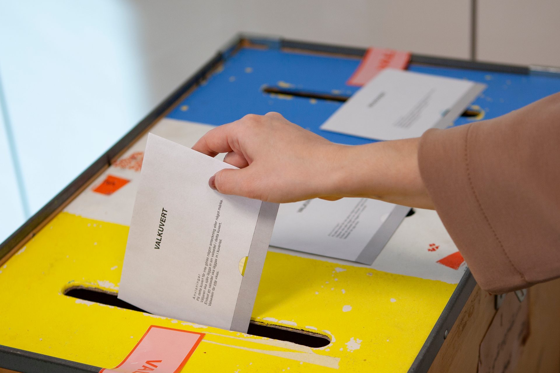 A hand drops a ballot envelope labeled "VALKUVERT" into a yellow and blue ballot box during an election.