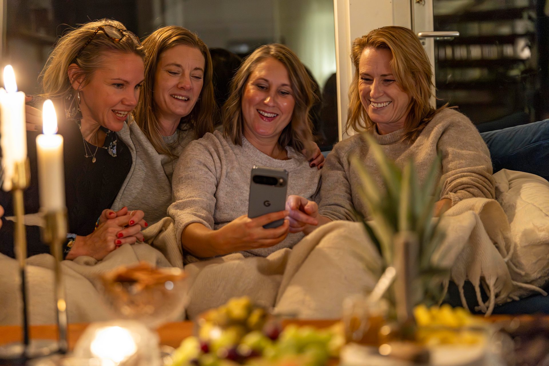 Four women laughing together at a phone in a cozy, candlelit room.