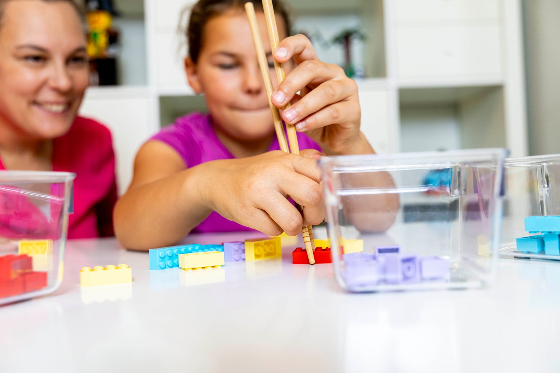 A child picks up colorful Lego bricks with chopsticks, watched by a smiling adult.
