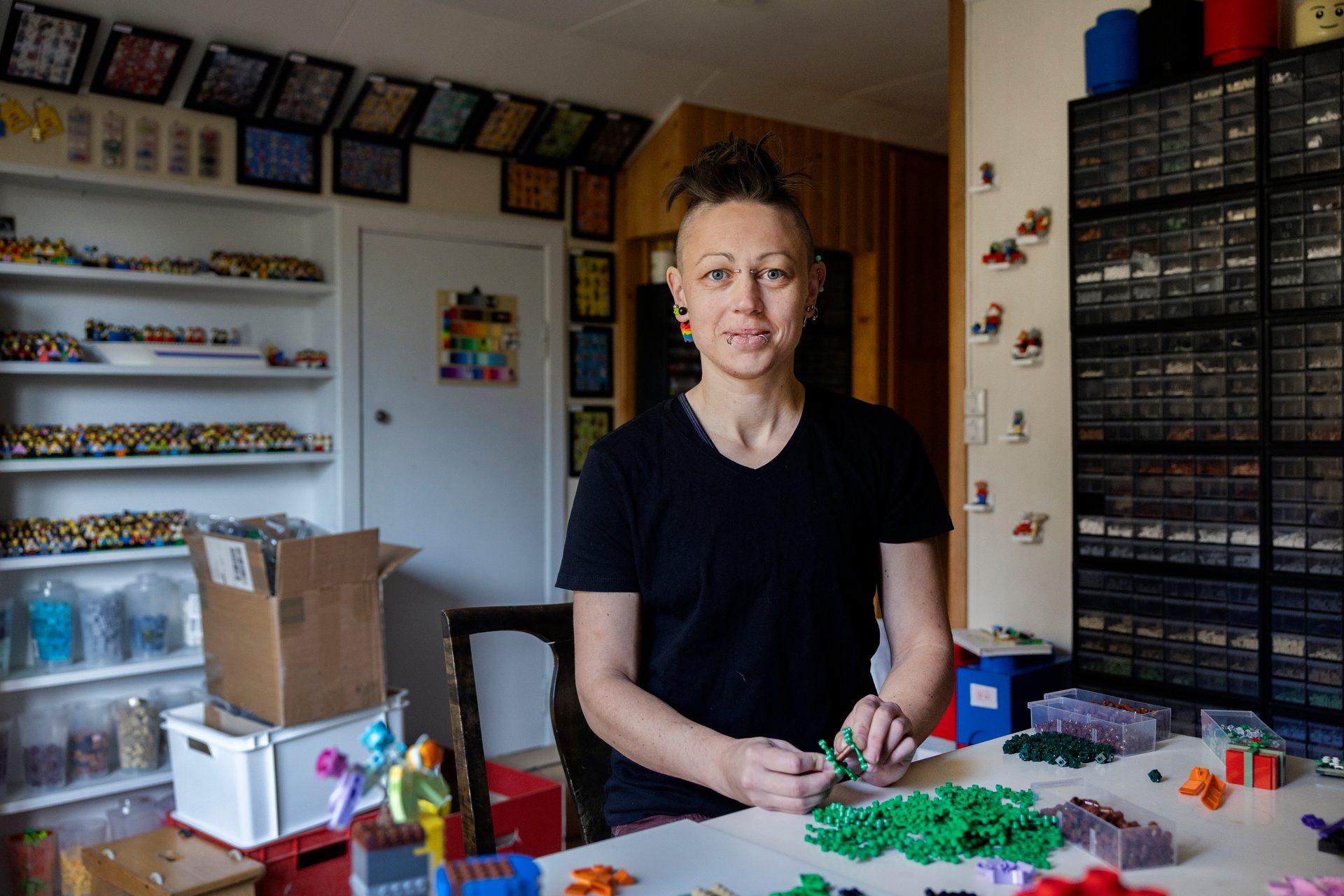 Person with unique hair and piercings smiles, holding green Lego pieces, surrounded by Lego collections.