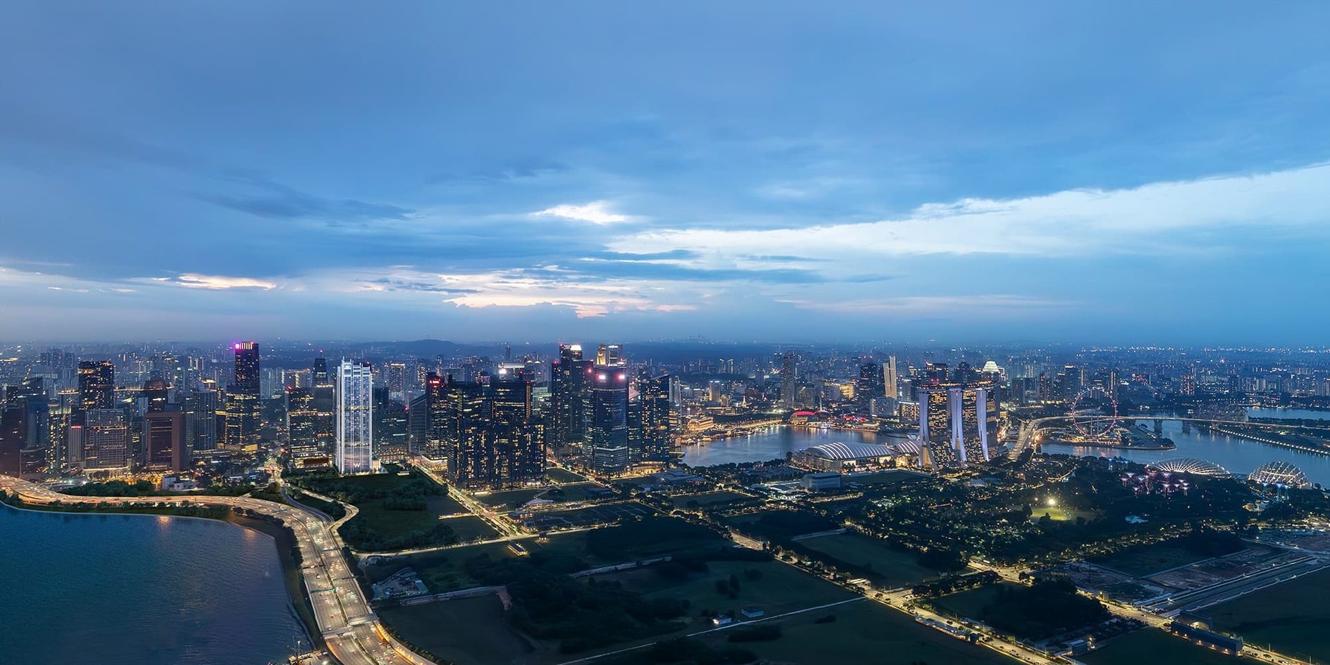Aerial night view of Singapore's illuminated cityscape, bay, and highways under a cloudy sky.