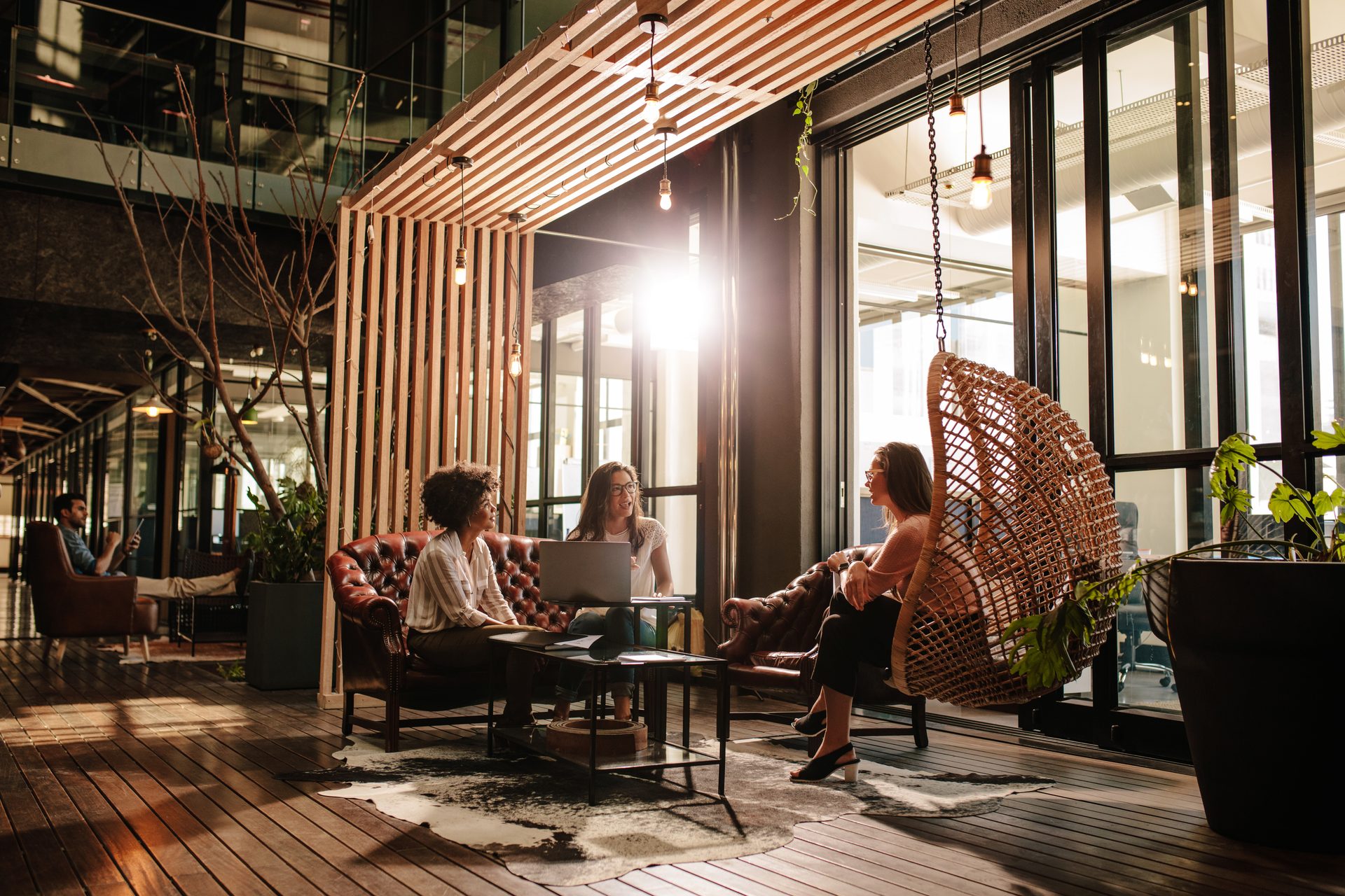 Three women collaborate in a stylish office lounge with a hanging chair and modern wooden decor.