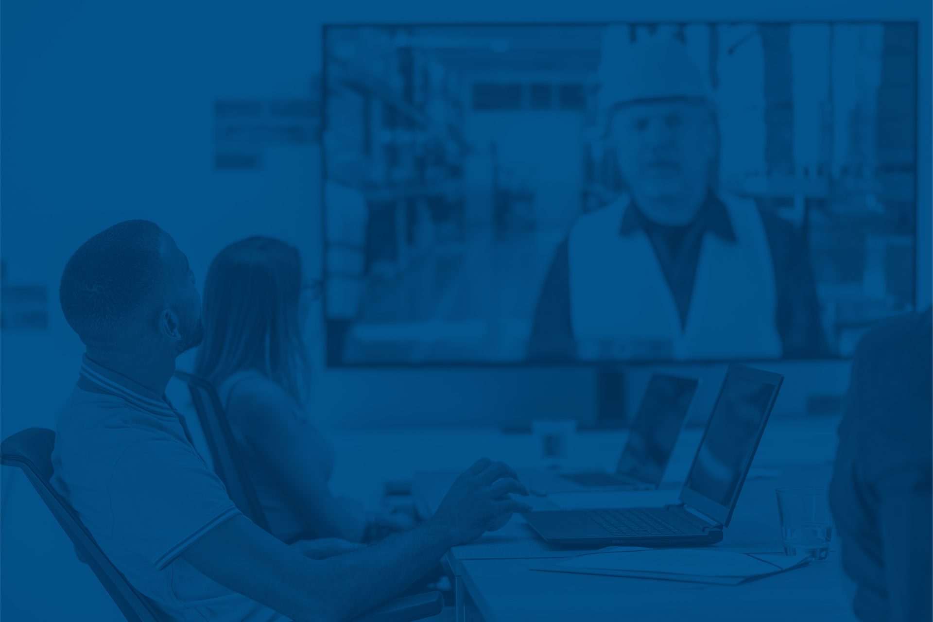Two people watch a virtual meeting with a man in a hard hat on screen.