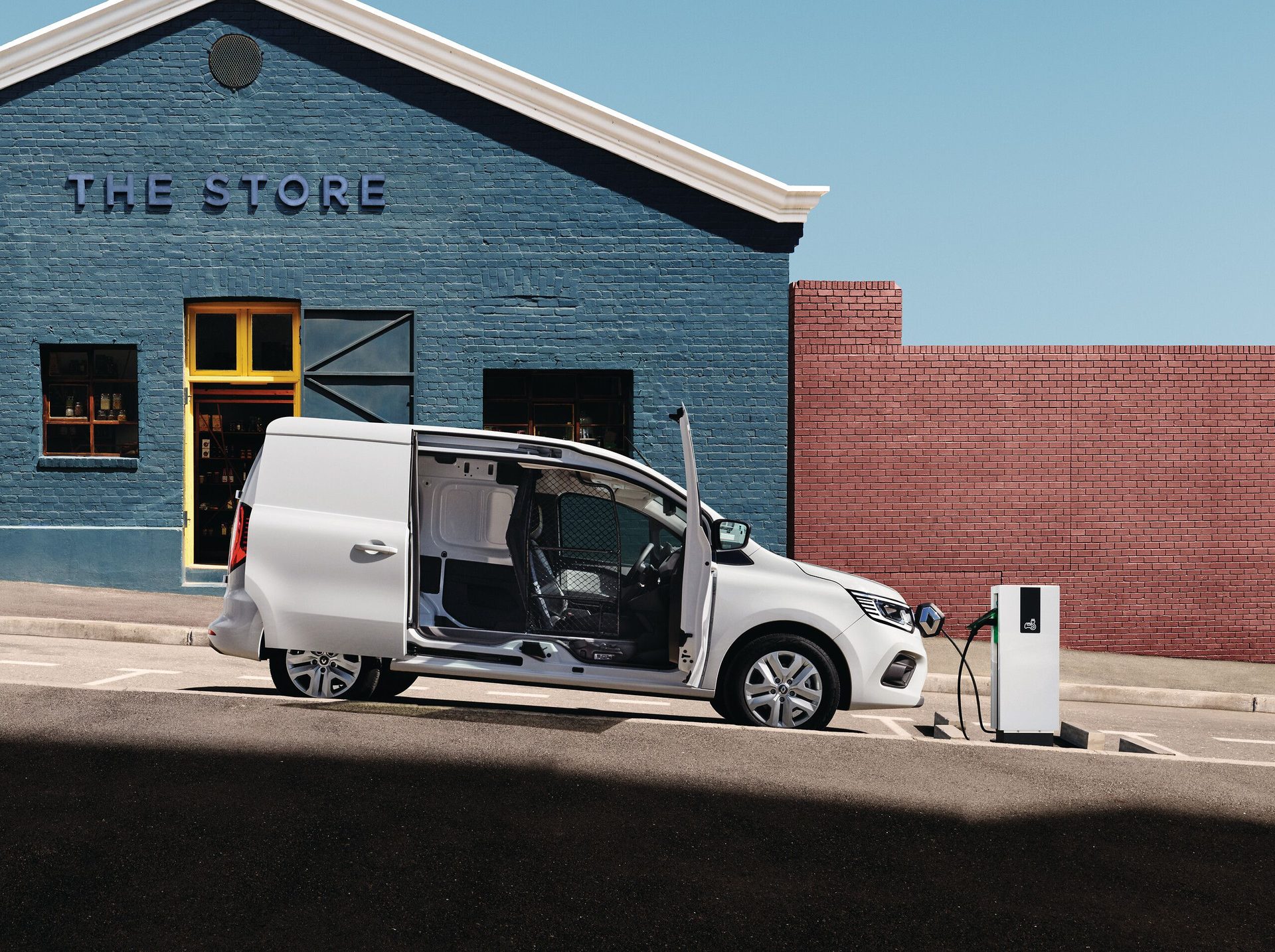 White electric delivery van with open doors charging outside "THE STORE" building on a sunny day.