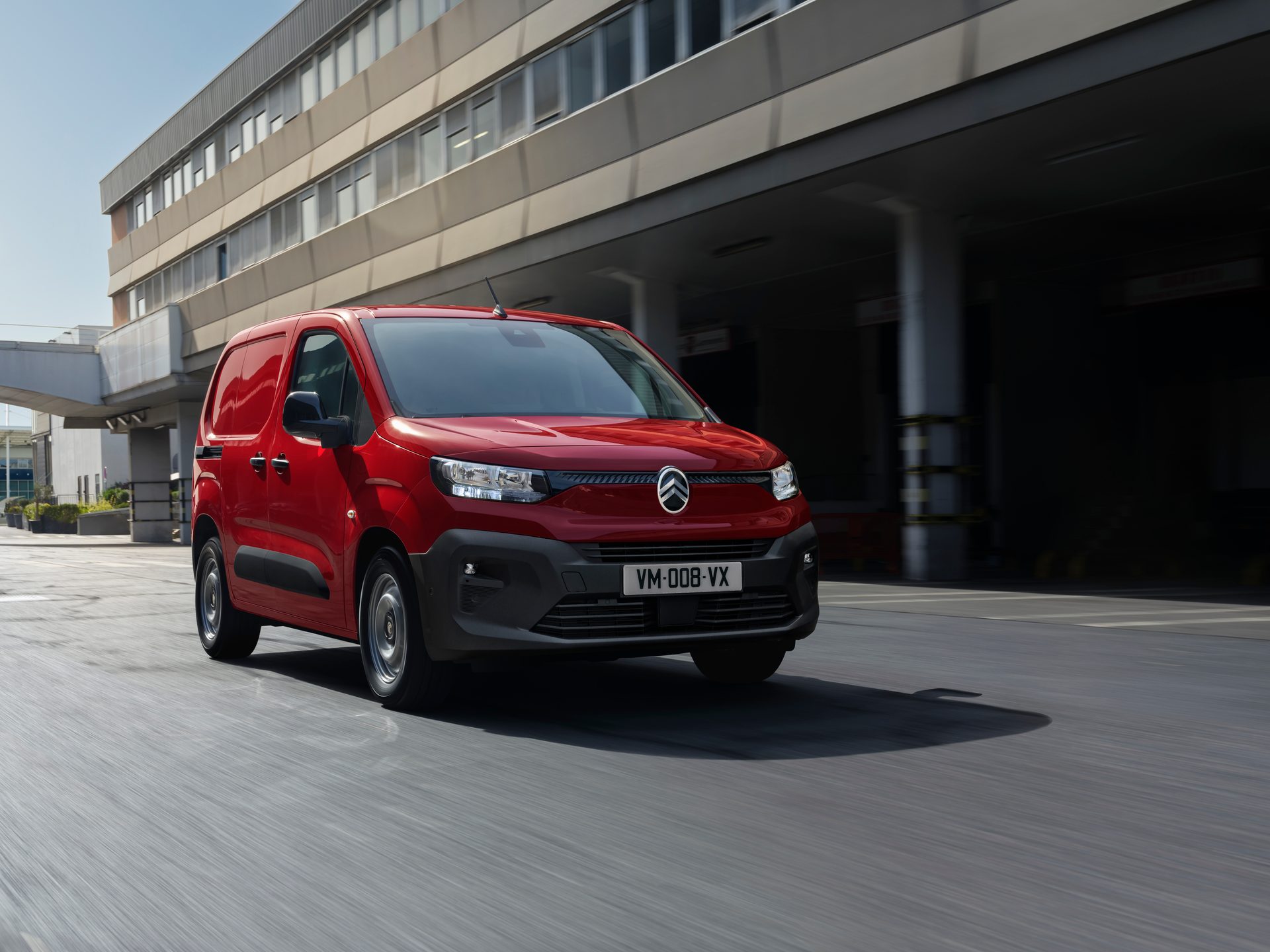 Red Citroen Berlingo van in motion on a road with a modern building in the background.
