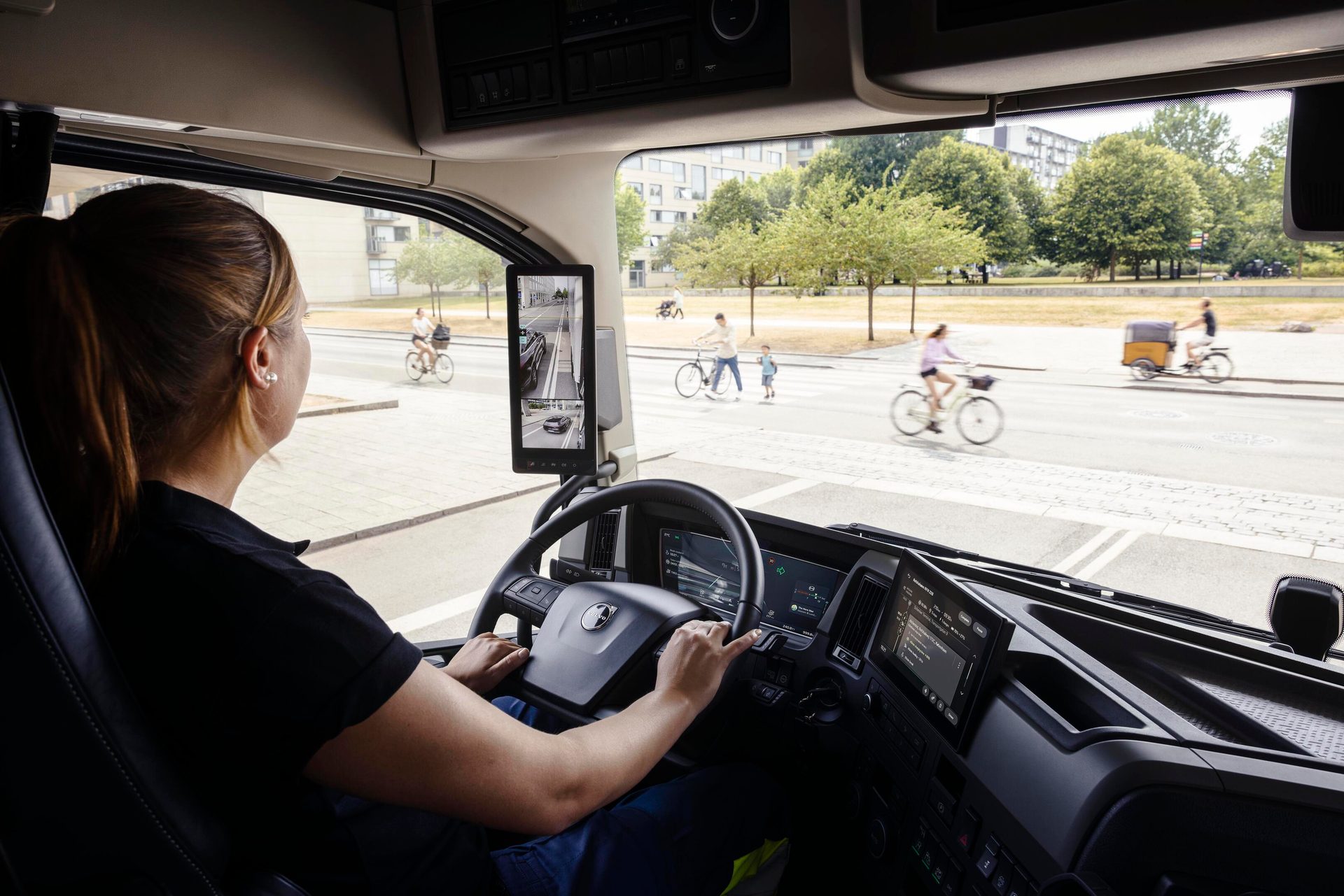 Driver in a truck looking at street traffic with cyclists and pedestrians, using a digital side mirror.