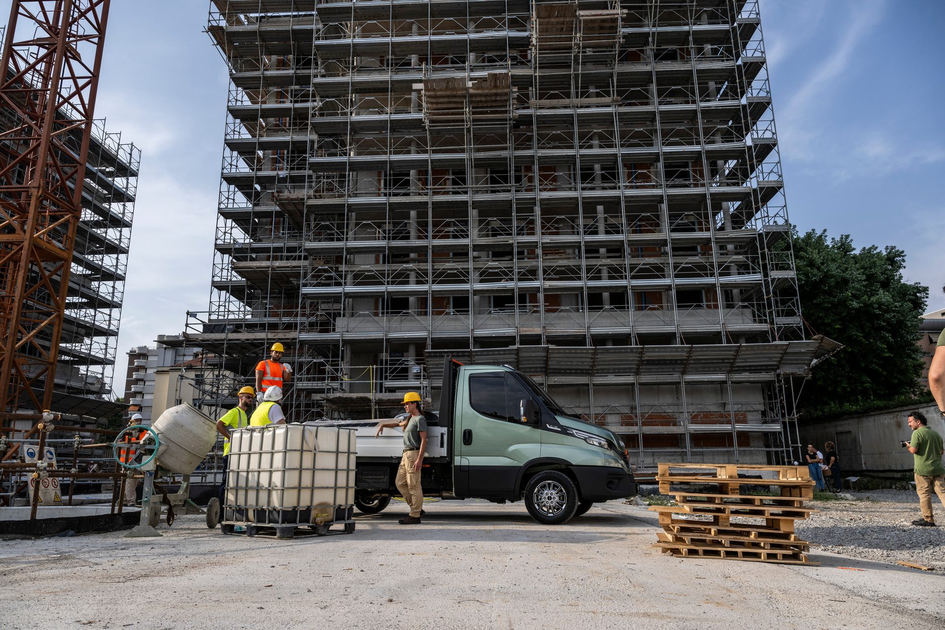Construction workers operate a concrete mixer and an IBC next to a green flatbed truck at a building site.