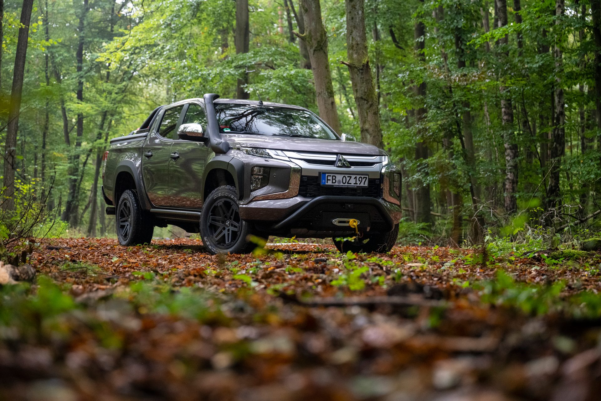 A gray Mitsubishi Triton pickup truck, equipped for off-road, parked on a leafy path in a dense forest.