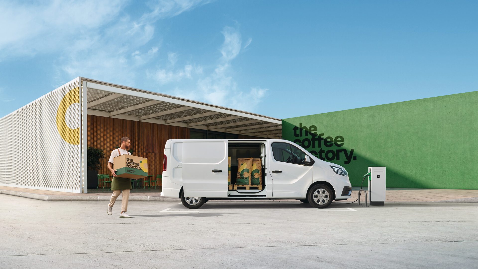 A man carries a coffee box from a charging electric van parked outside a modern "the coffee factory" building.