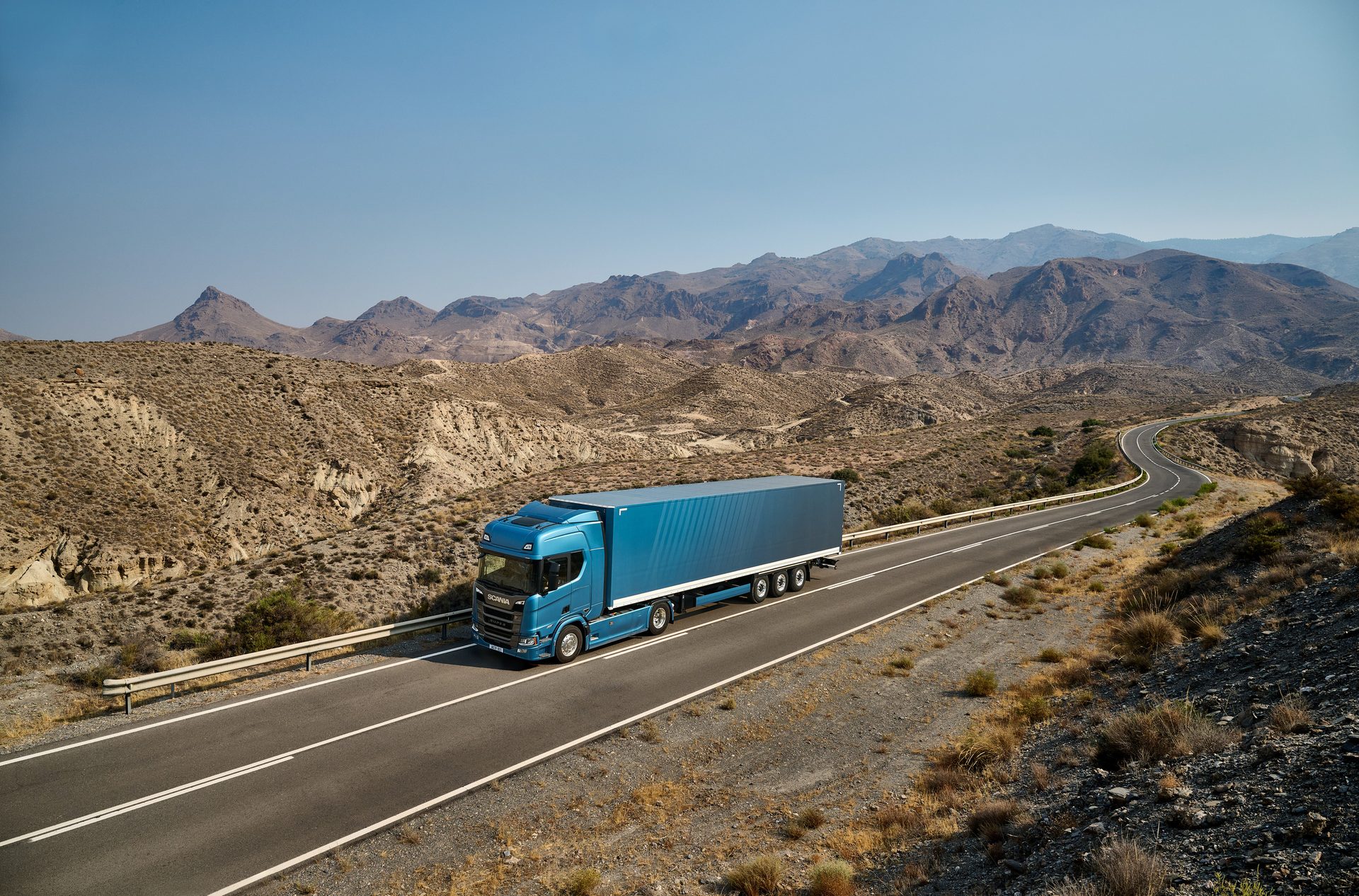 Blue semi-truck with trailer drives on a winding road through a dry, mountainous desert landscape.
