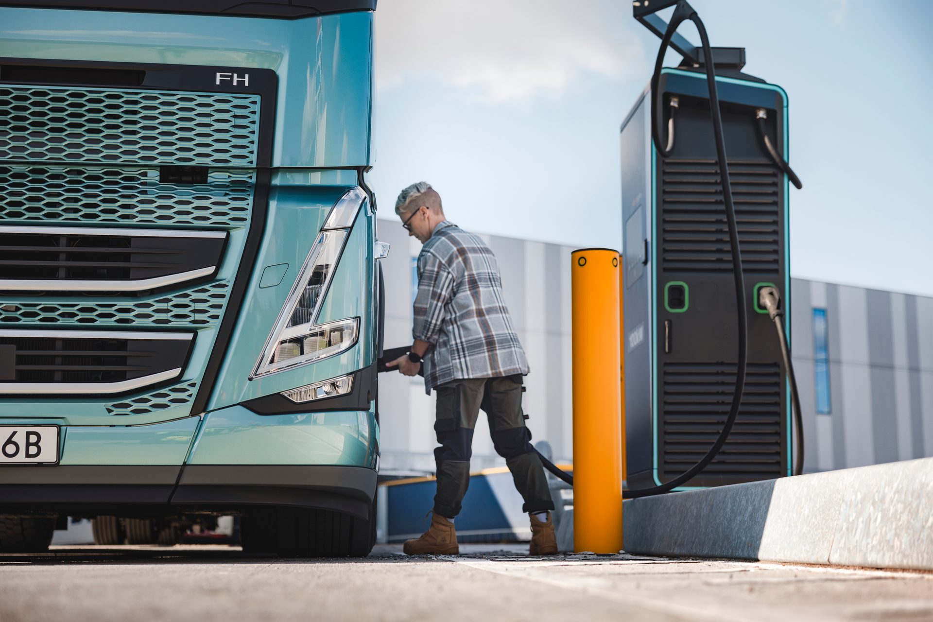 A person is charging a large teal electric truck at an outdoor charging station under a clear sky.