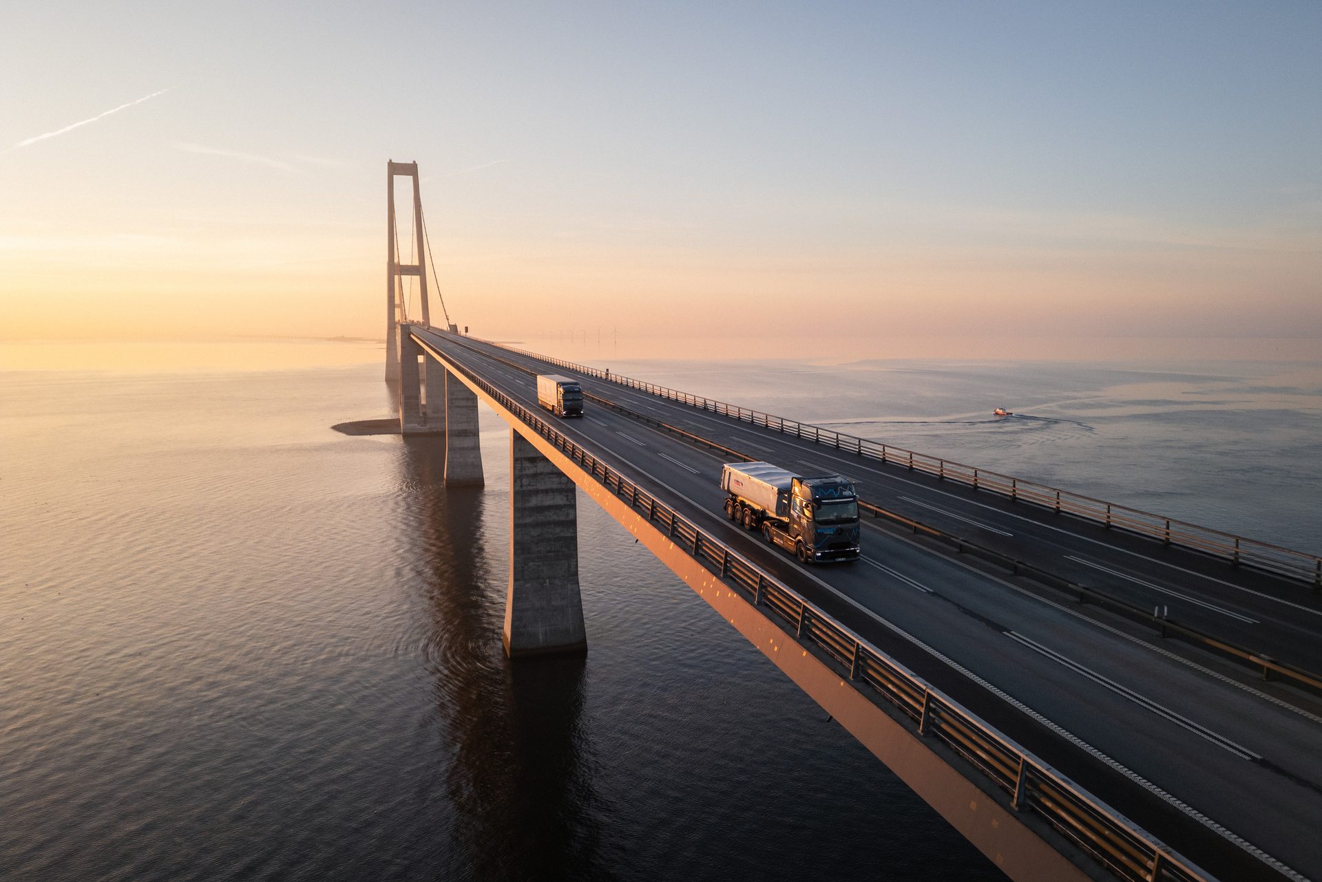 Aerial view of two trucks driving on a long suspension bridge over water at sunrise or sunset.