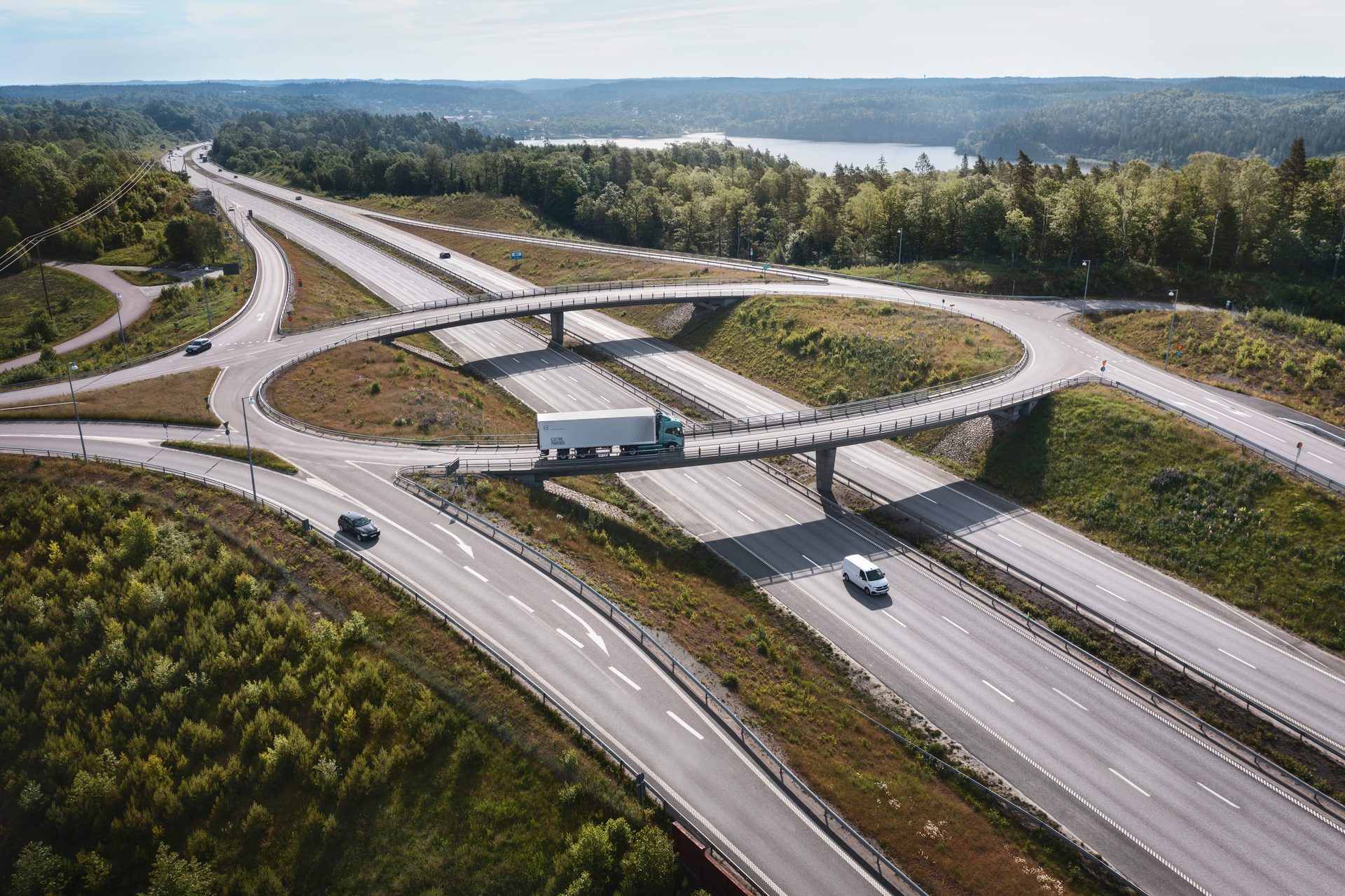 Aerial view of a complex highway interchange with a truck and cars, surrounded by forests and a distant lake.
