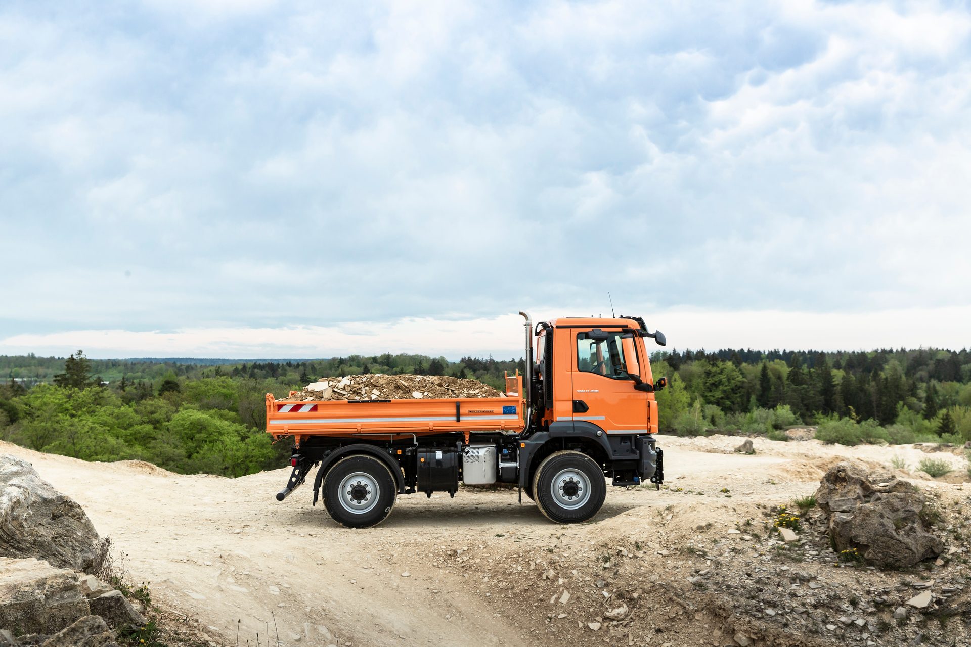 Orange dump truck with debris on a dirt road in a quarry, forest in background, cloudy sky.