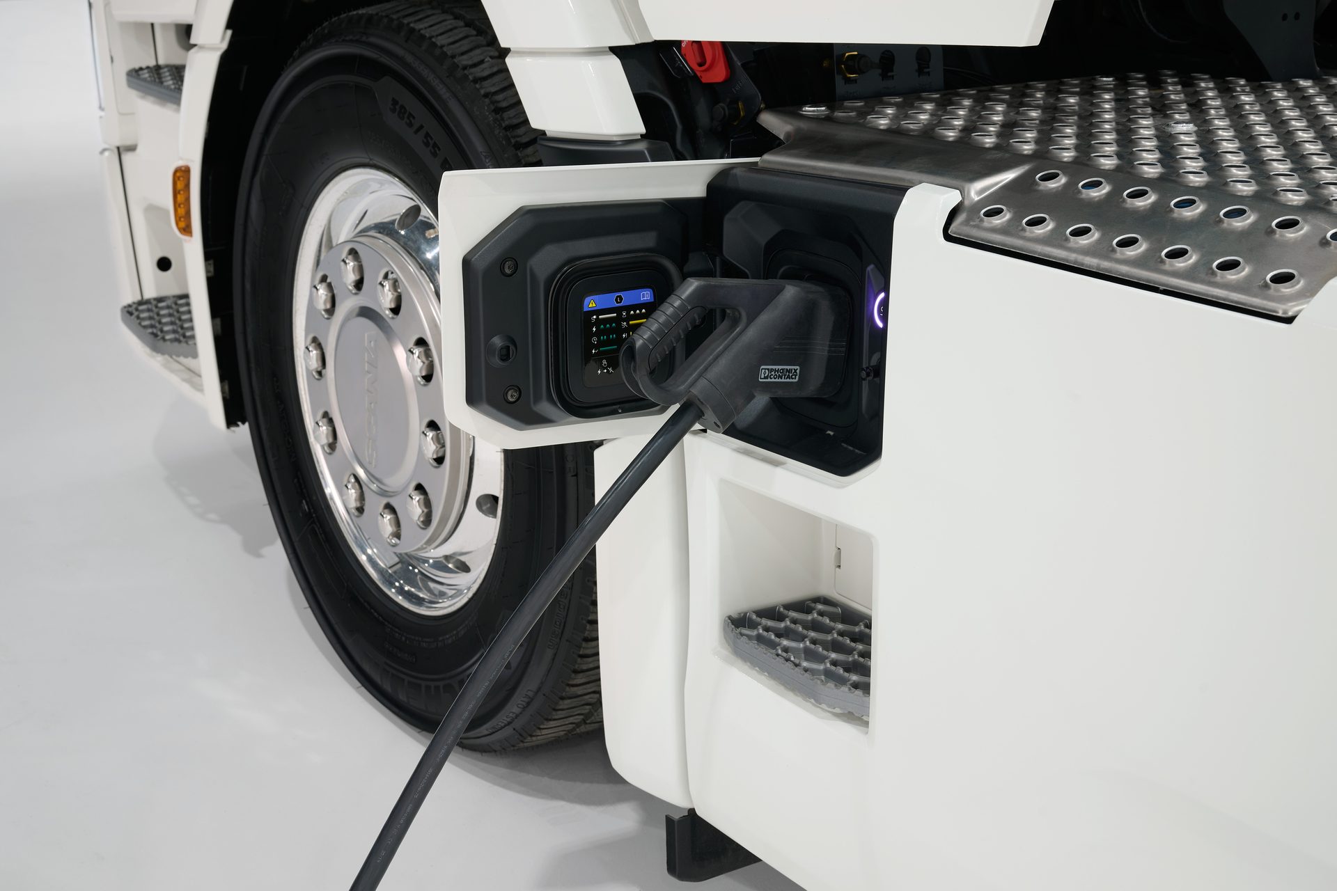 Electric truck charging port with cable plugged in, next to a large wheel and a metal step.