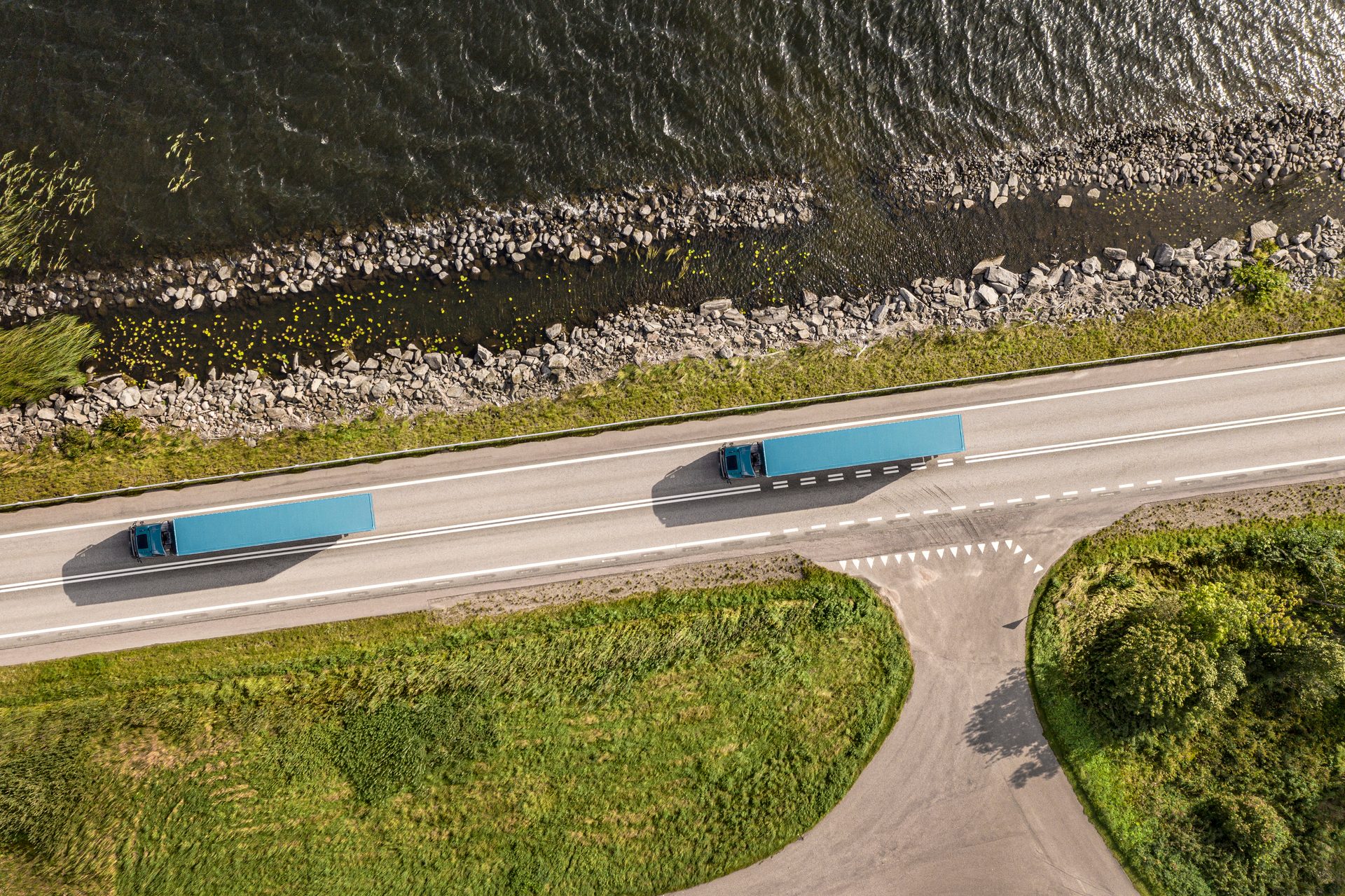 Aerial view of two blue semi-trucks driving on a road alongside a body of water and green land.