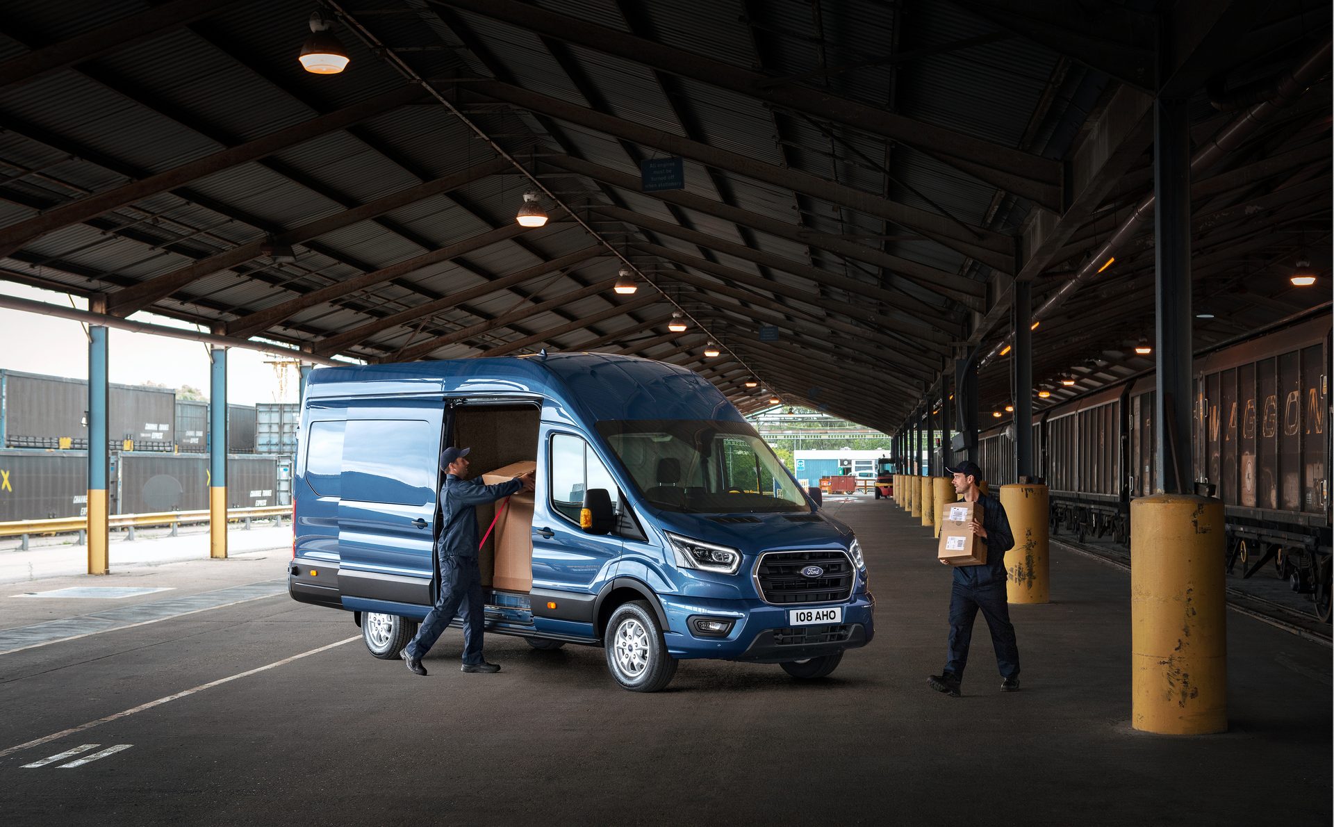 Two delivery workers load and unload packages from a blue Ford Transit van at a busy cargo depot.