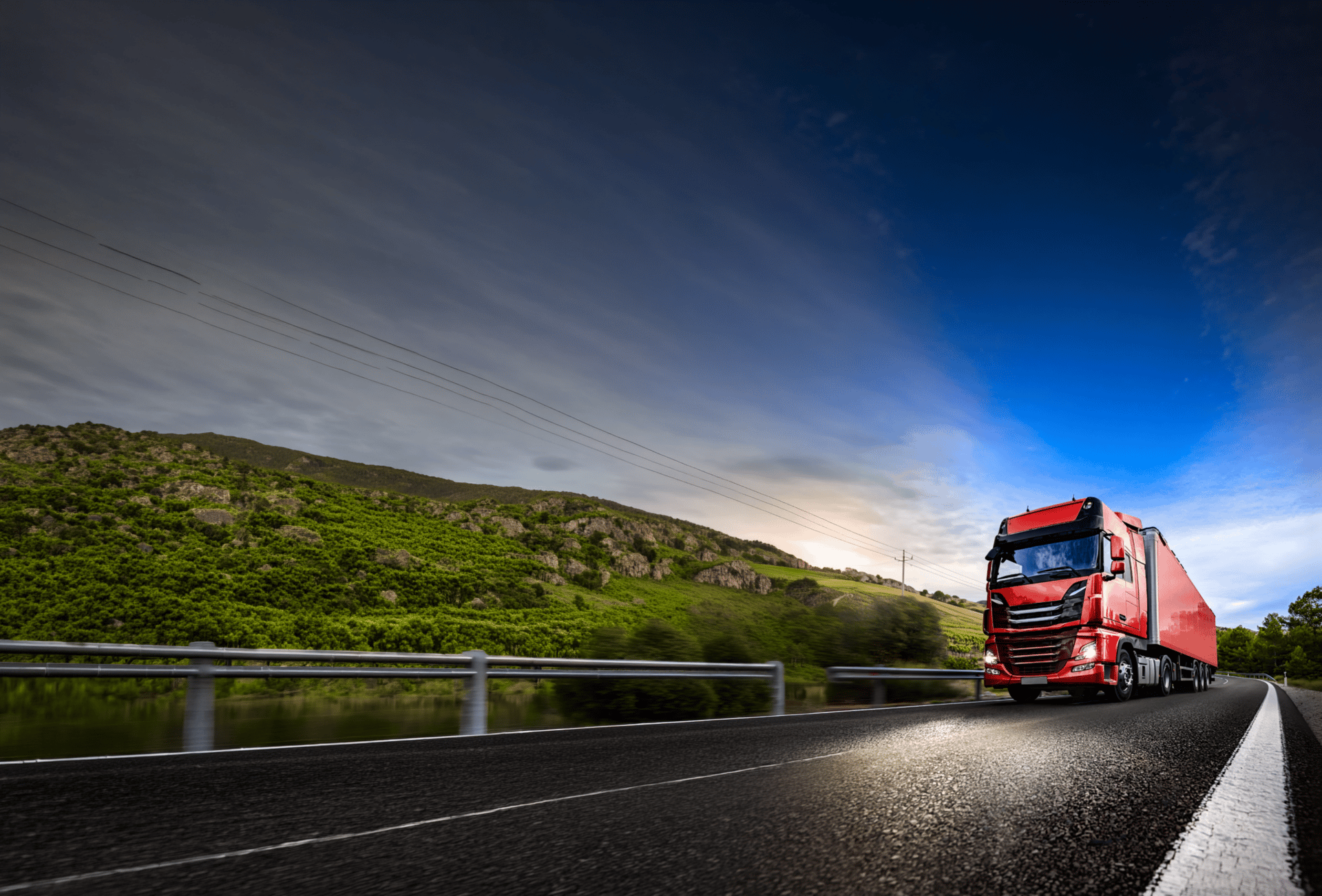 A red semi-truck drives on a winding road next to a river with green hills under a bright blue sky.