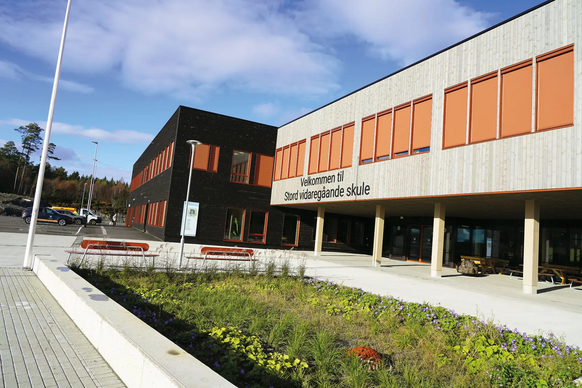 Modern school building with dark and light wood facades, a welcoming sign, and surrounding landscaping.