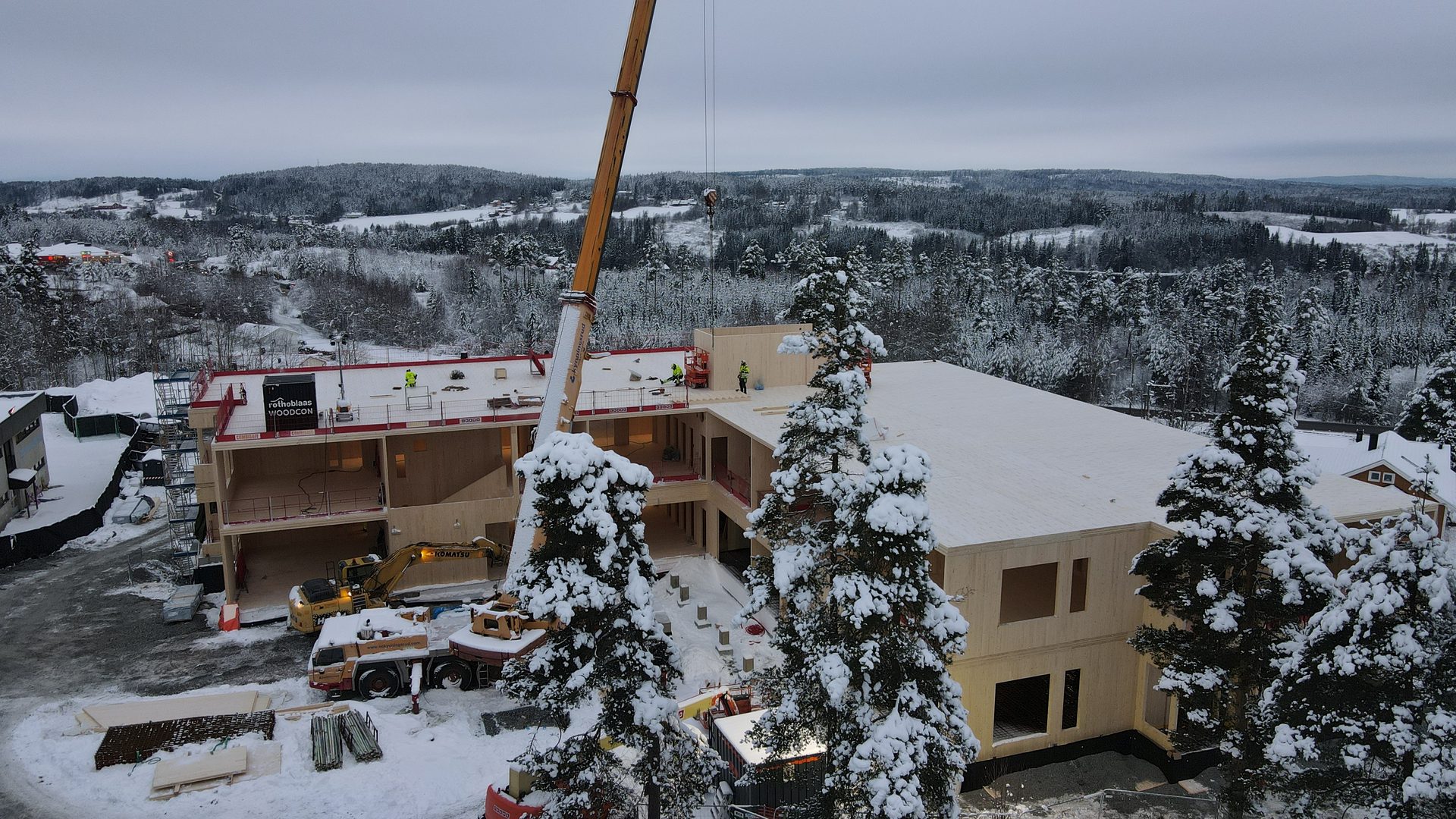Large wooden building under construction in a snowy forest, with a crane and workers.
