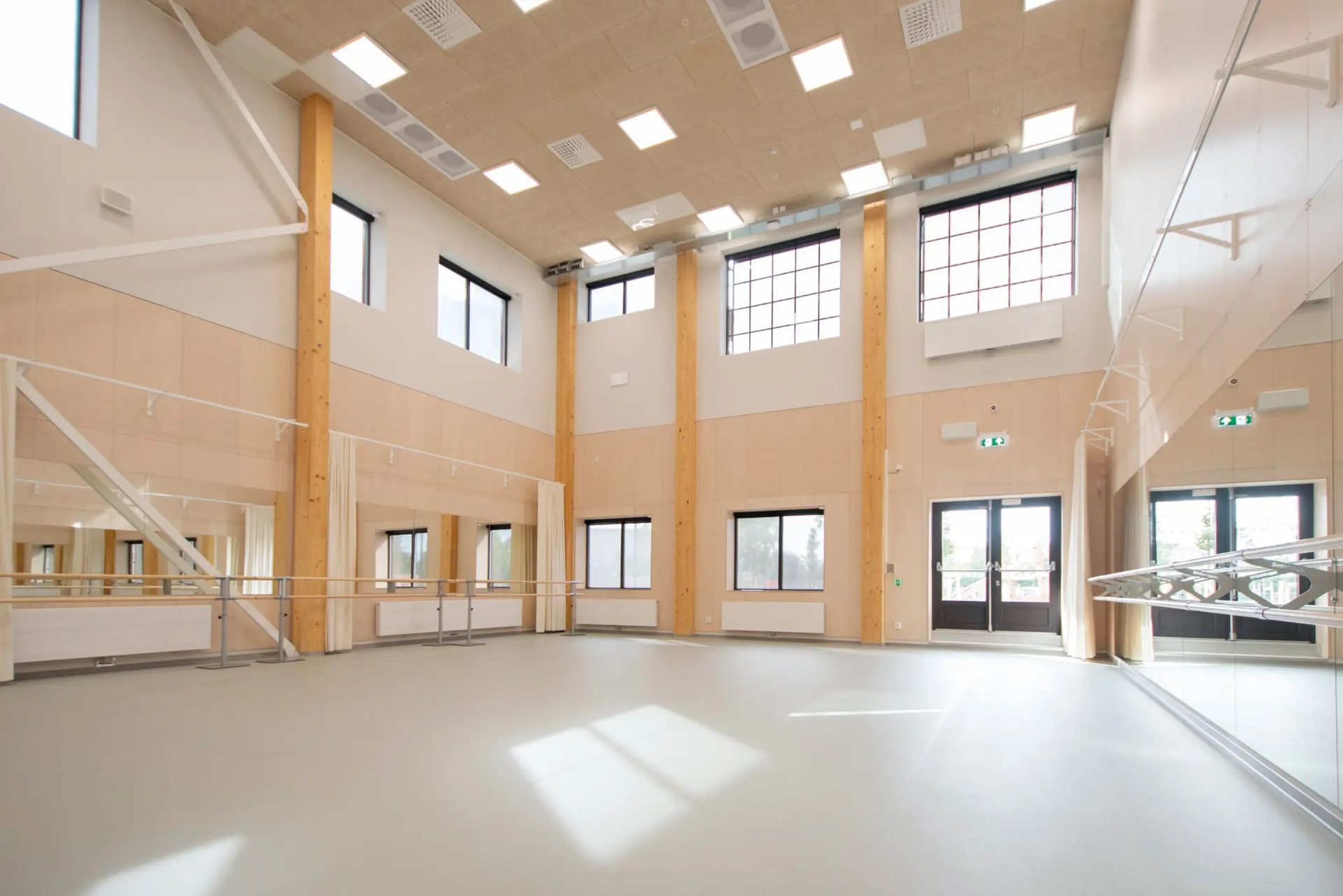 Bright, modern dance studio with wooden walls, tall windows, mirrored walls, ballet barres, and high ceilings.
