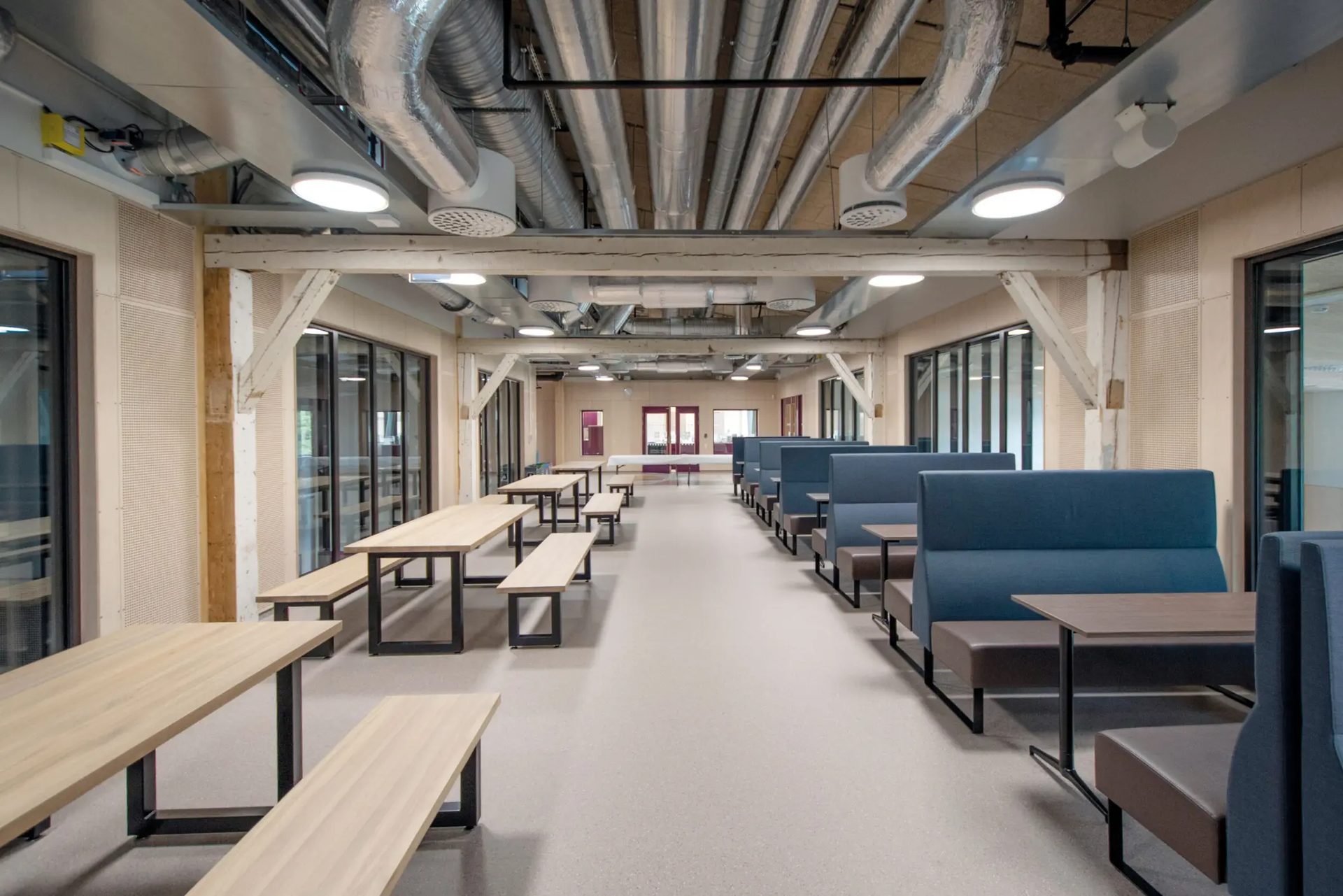 A long, modern cafeteria with tables, benches, and blue booths, exposed ceiling, and wooden beams.